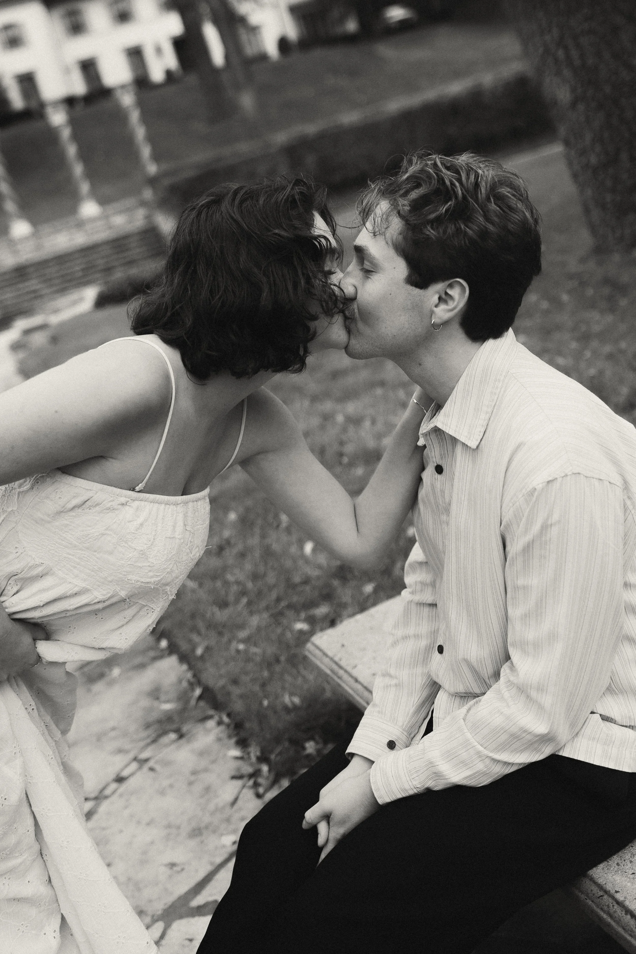 A black-and-white photo of a woman and a man kissing outdoors, with the woman leaning forward and the man sitting on a park bench.