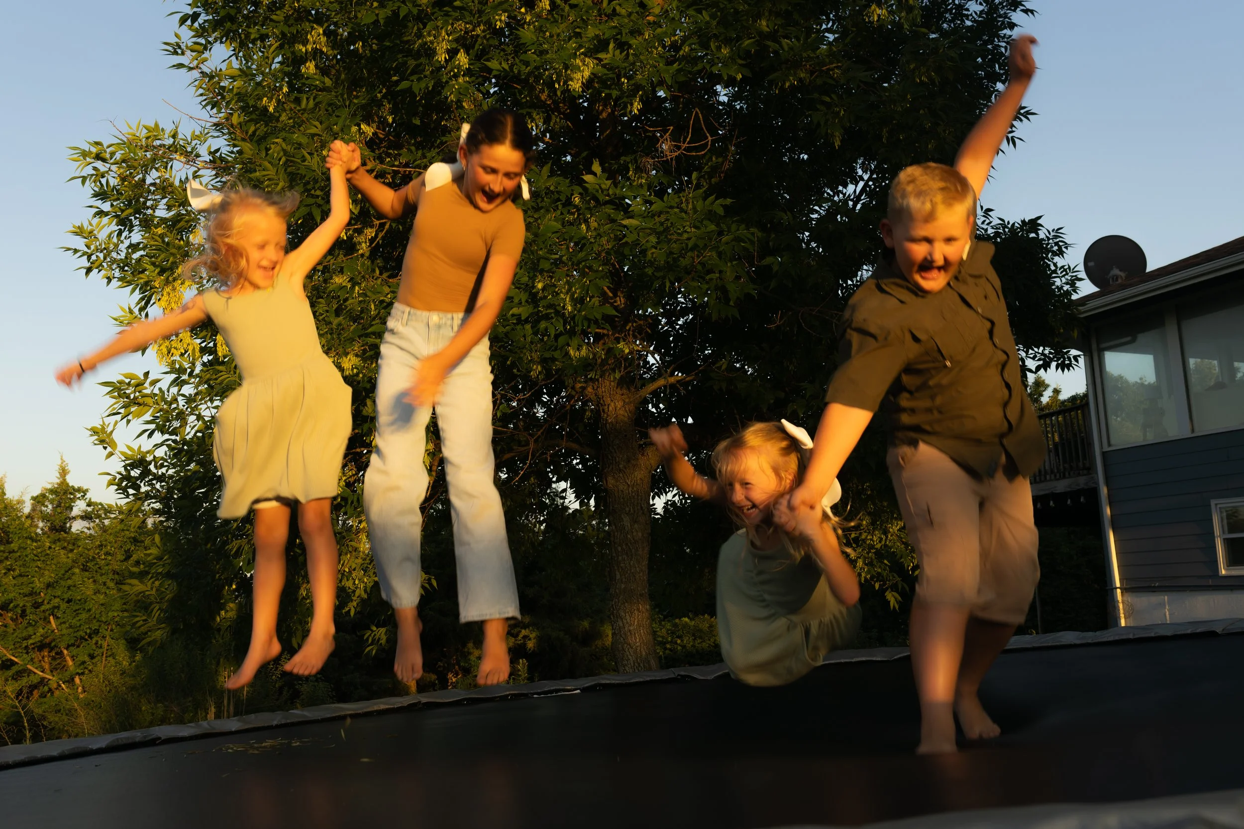 Children jumping on a trampoline outdoors during sunset, with trees and a house in the background.