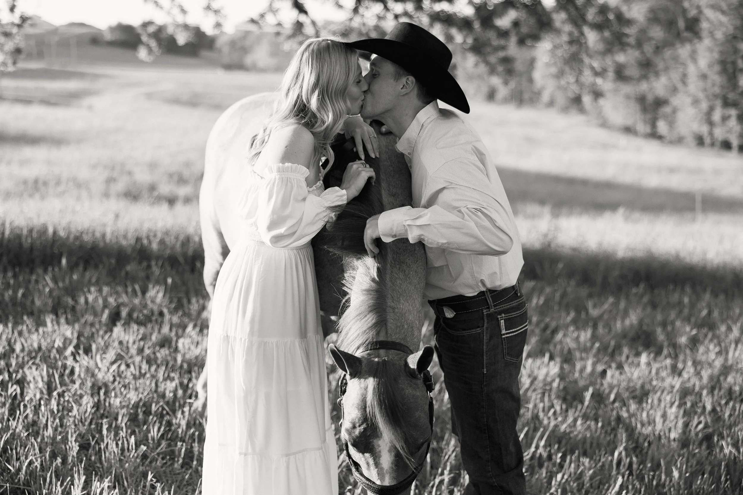 A man wearing a cowboy hat and a woman in a white dress share a kiss while holding a horse's head in an open field.