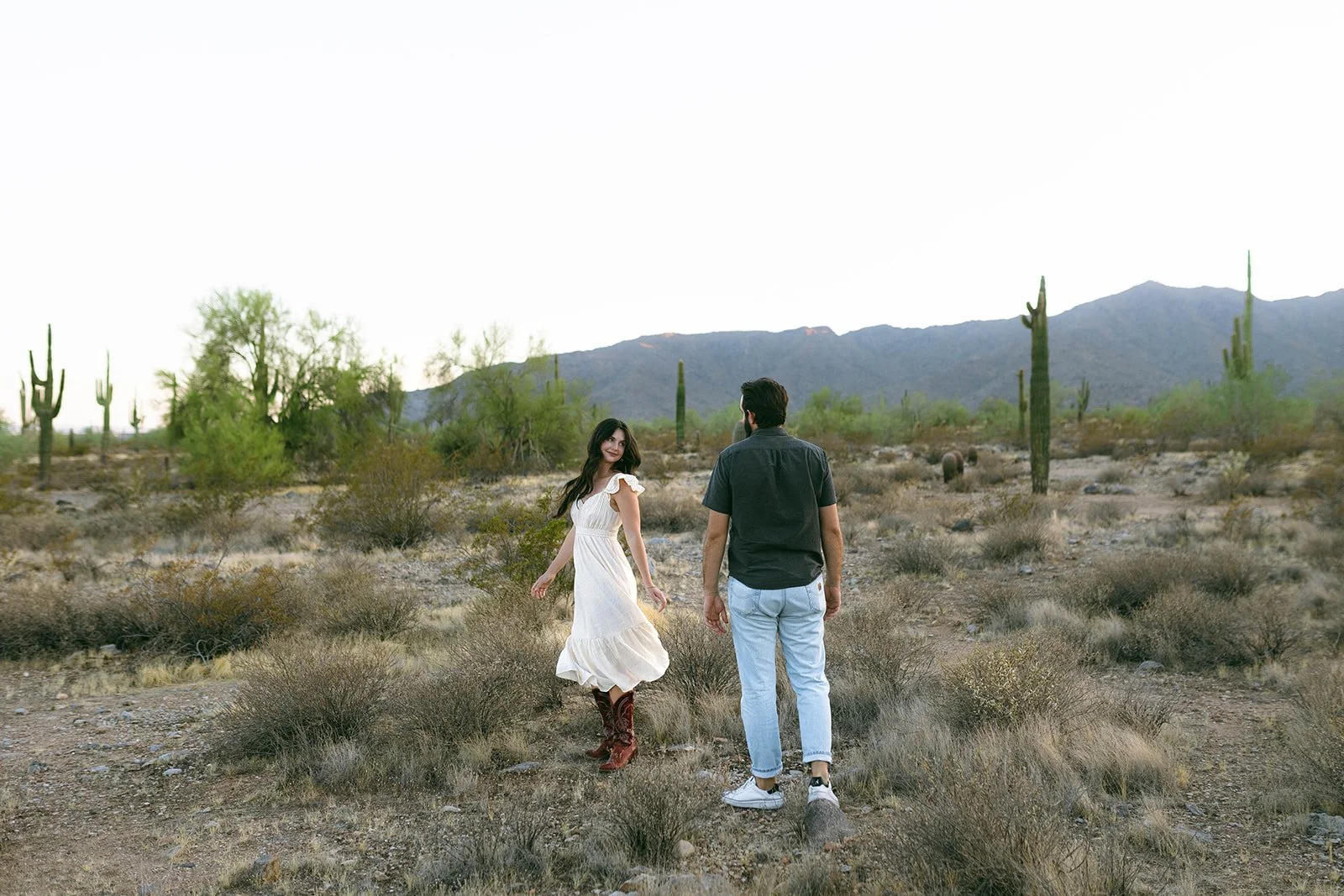 A man and woman walking in a desert landscape with cacti, mountains in the background, and a clear sky.