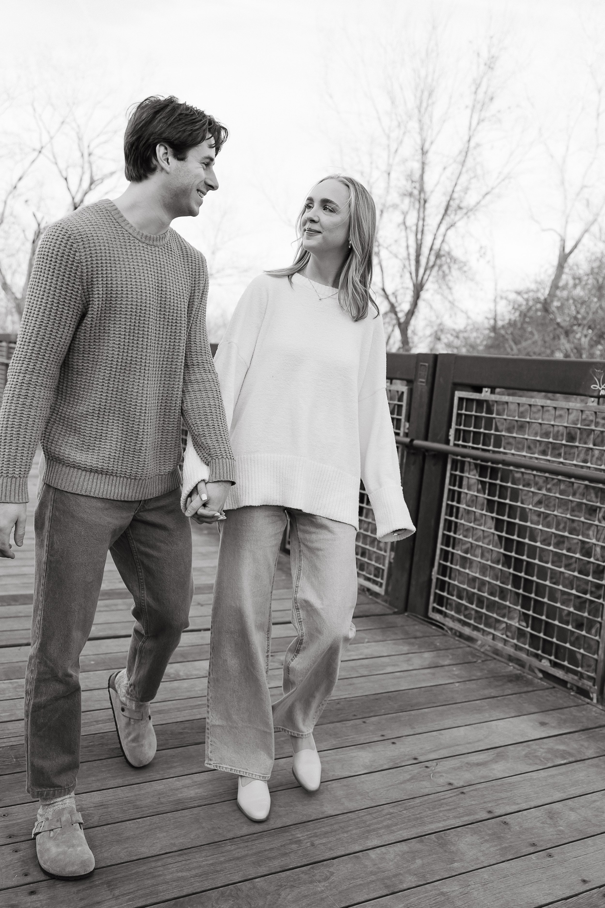 A black-and-white photo of a young couple holding hands and walking on a wooden bridge outdoors, smiling at each other, with bare trees in the background.