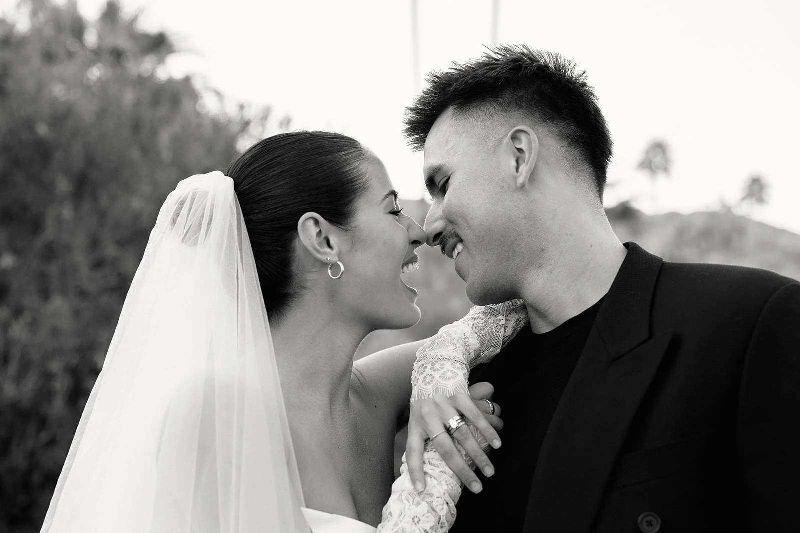 A black-and-white photo of a bride and groom on their wedding day, smiling and touching their noses together, outdoors with palm trees in the background, in Palm Springs, California.