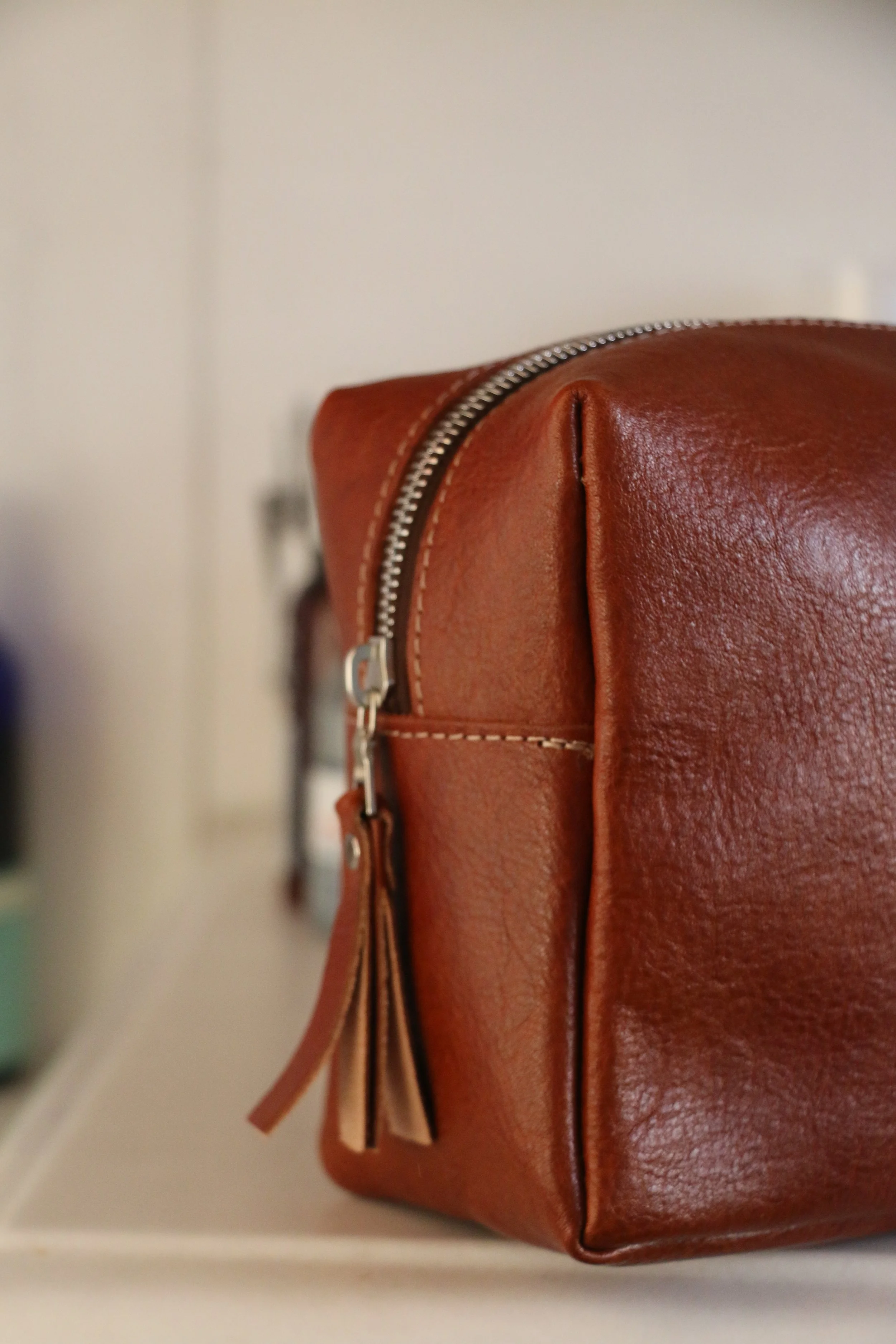 Close-up of a brown leather bag with a visible zipper and tassel, against a neutral background.