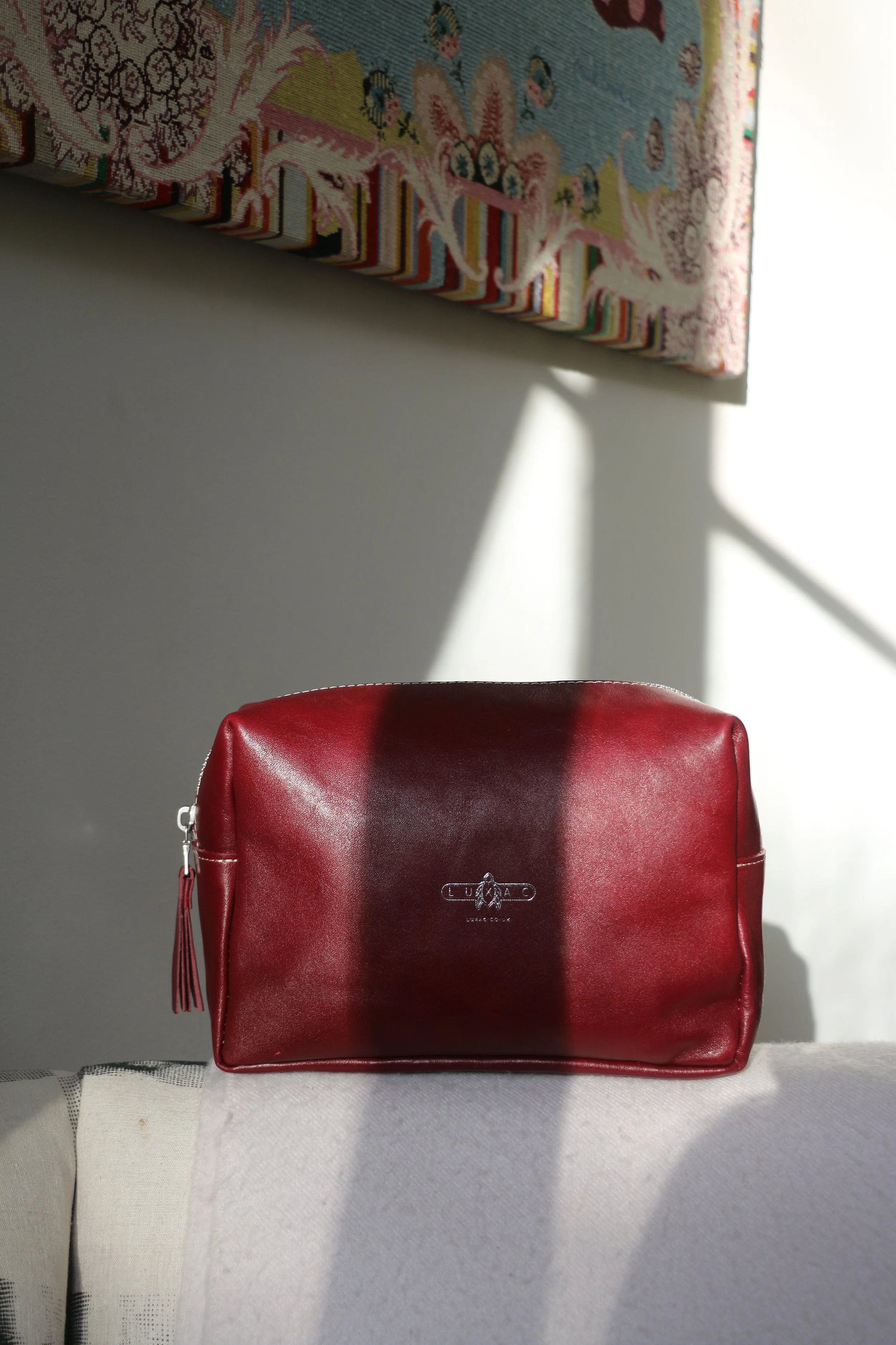 A red leather bag with a tassel zipper, placed on a white surface. In the background, sunlight casts shadows on a white wall and a colorful tapestry with intricate patterns.