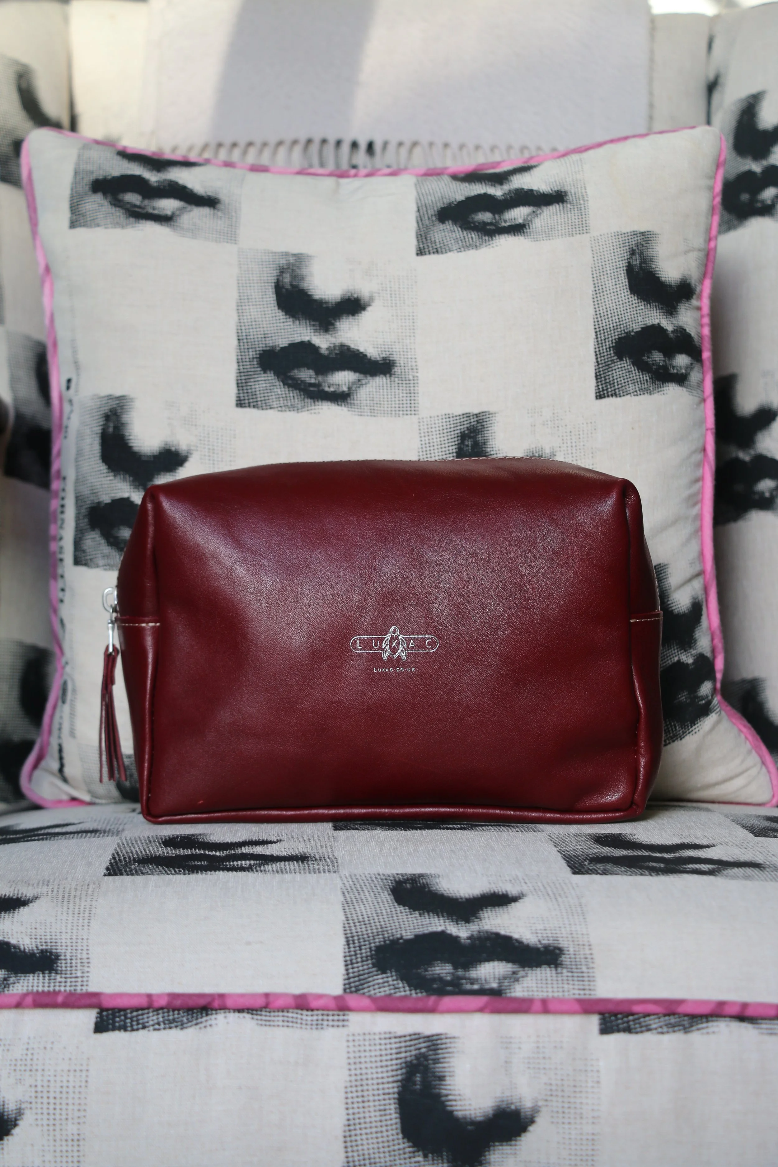 A maroon leather clutch purse with a small logo, placed on a decorative pillow featuring black and white print of women's faces.