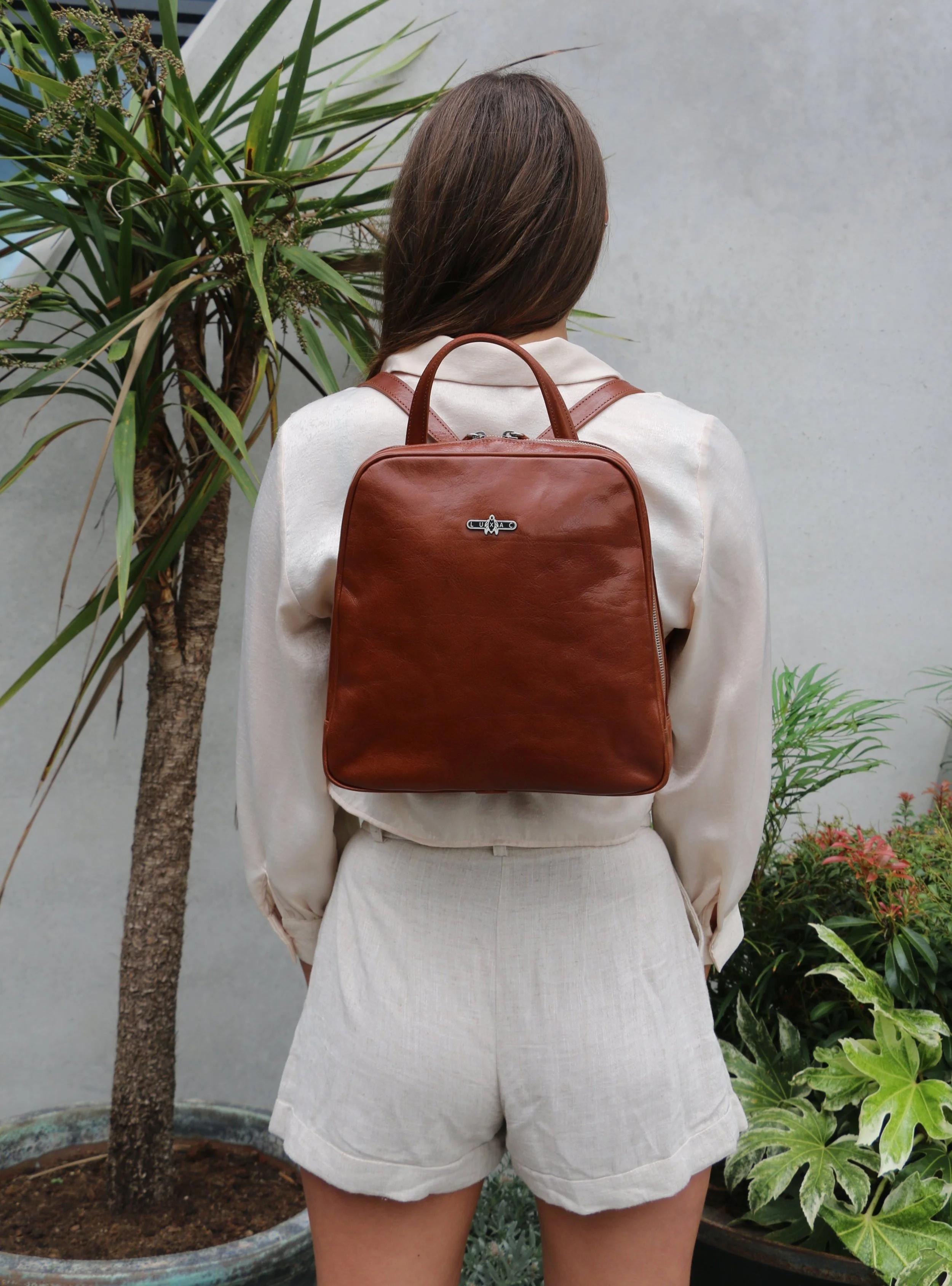 Back view of a woman with long brown hair wearing a cream blazer and shorts, carrying a brown leather backpack, standing next to a potted plant with green leaves.