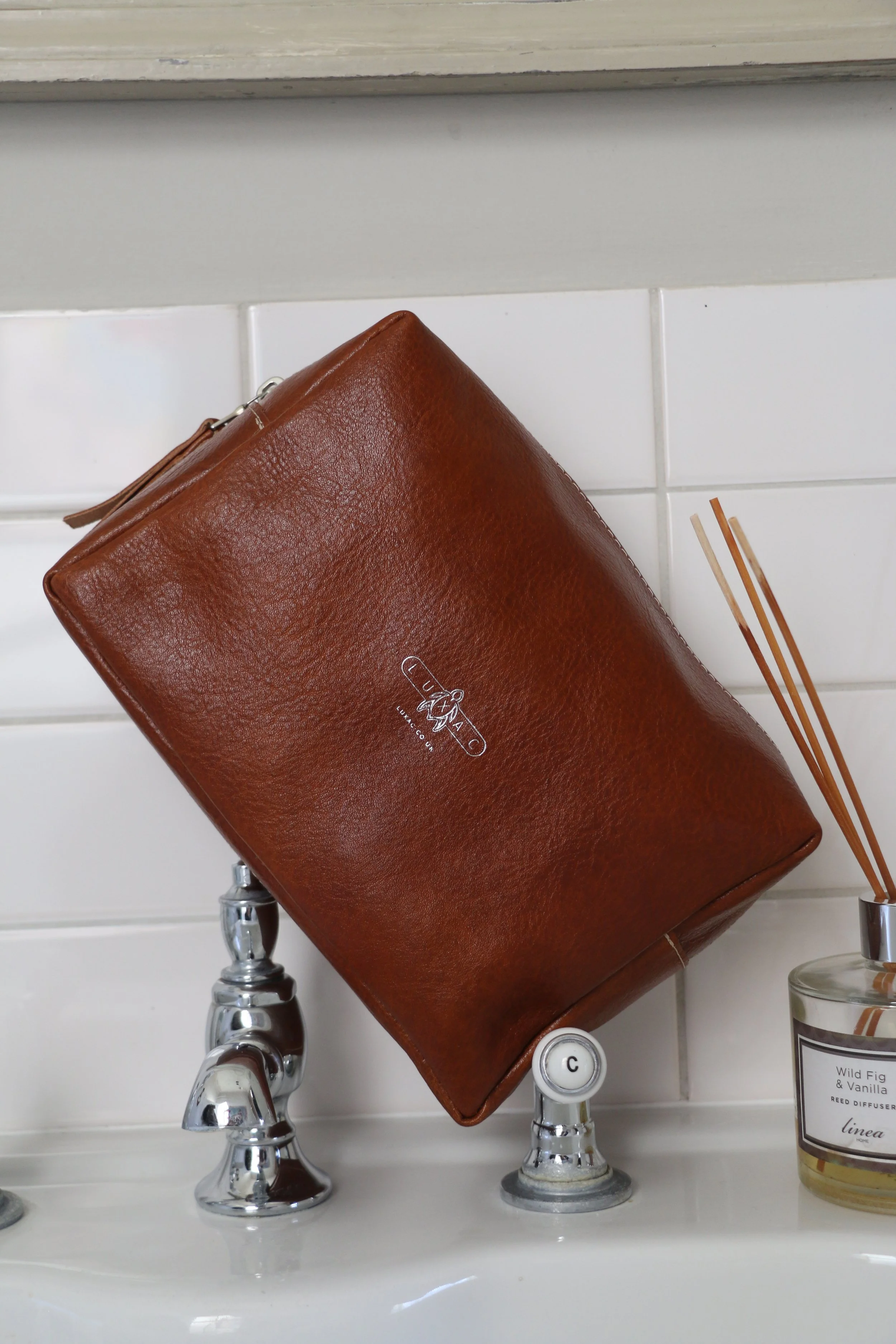 A brown leather GUESS clutch purse placed upside down on bathroom sink, with chrome faucet handles and a reed diffuser nearby.