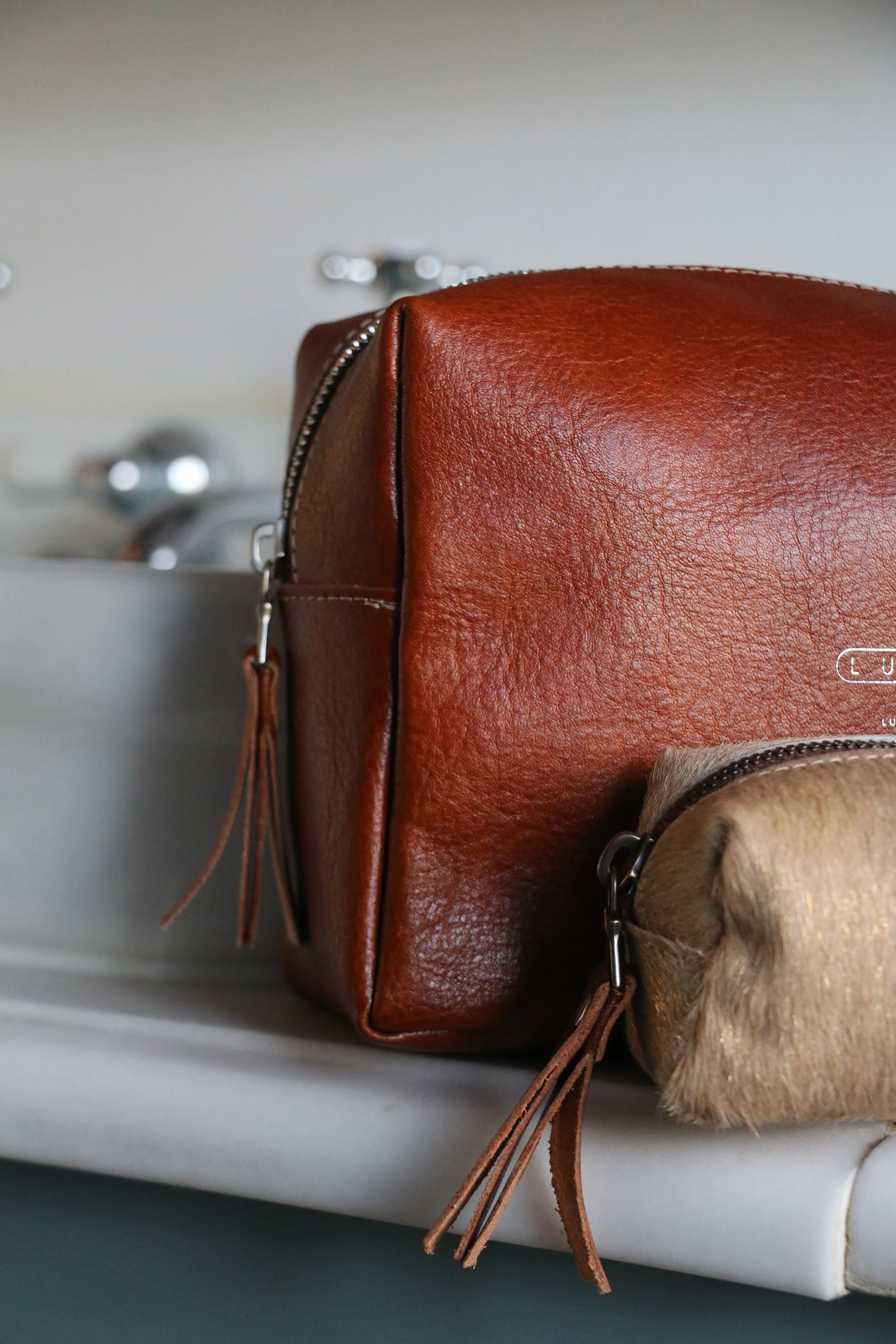 Close-up of a brown leather bag with tassel zipper pulls resting on a ledge near a sink.