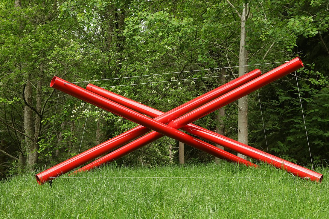 Two large red metal pipes crossed in front of a green forest background, supported by thin wires.