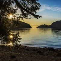 A peaceful lakeside scene at sunset with calm water, surrounded by trees and hills in the background.