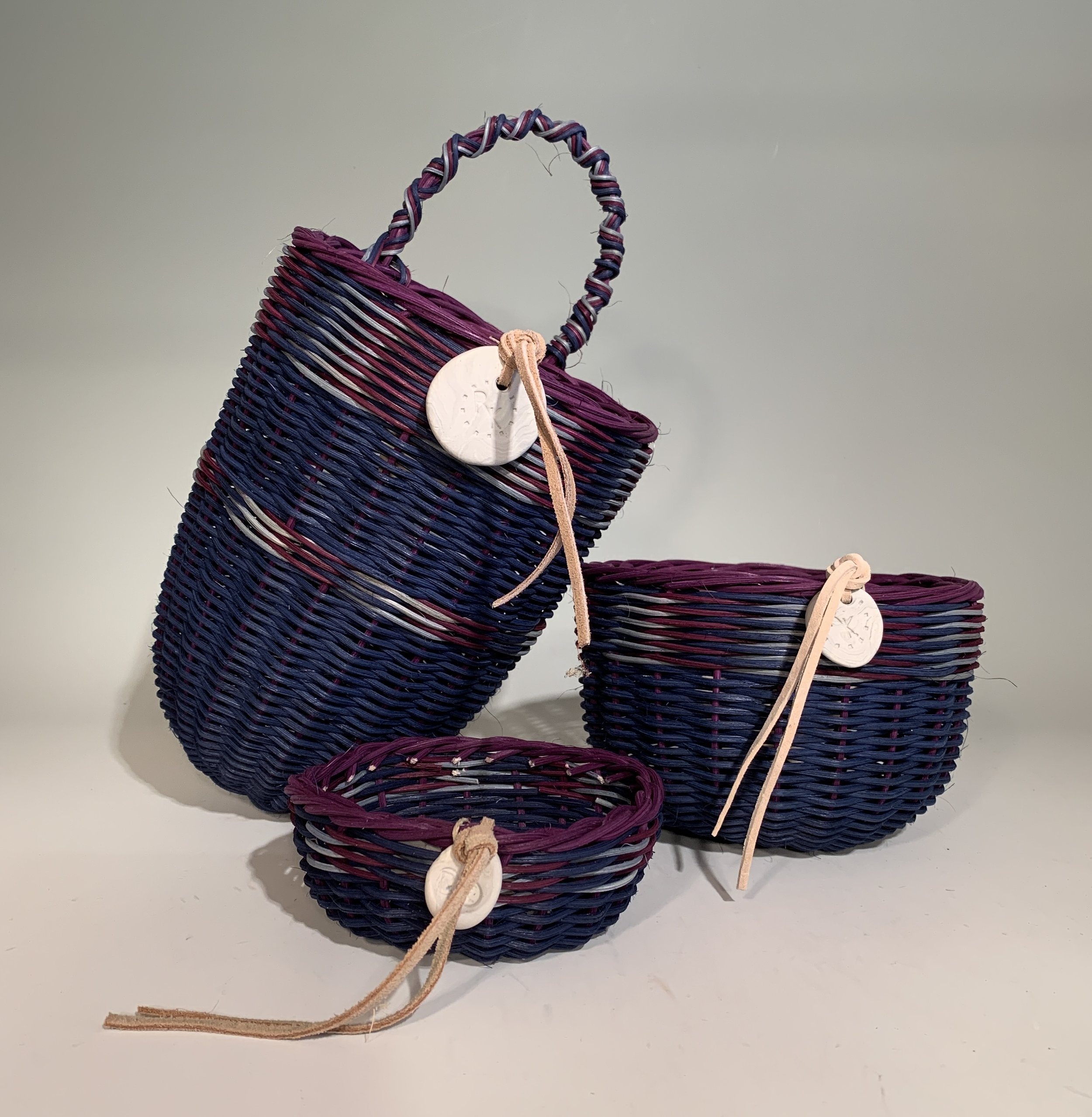 Three woven baskets with leather straps and tags, in different sizes and colors, arranged on a white surface against a plain background.