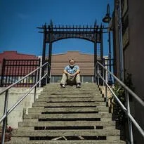 A person sitting on the steps of an outdoor staircase with metal handrails, leading up to an iron gate and a brick building in the background, under a clear blue sky.