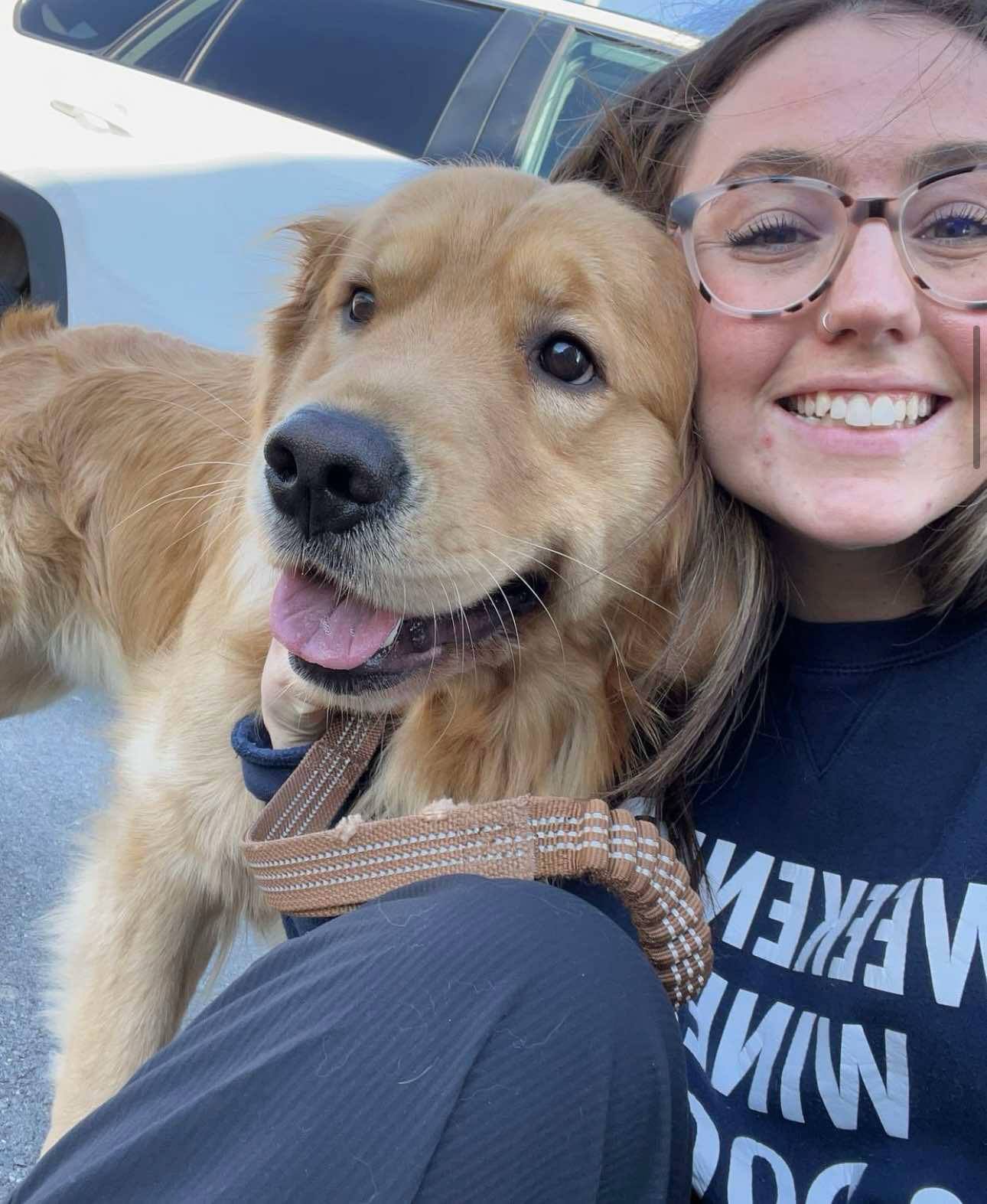 A person smiling next to a golden retriever dog with a leash, both taking a selfie outdoors.