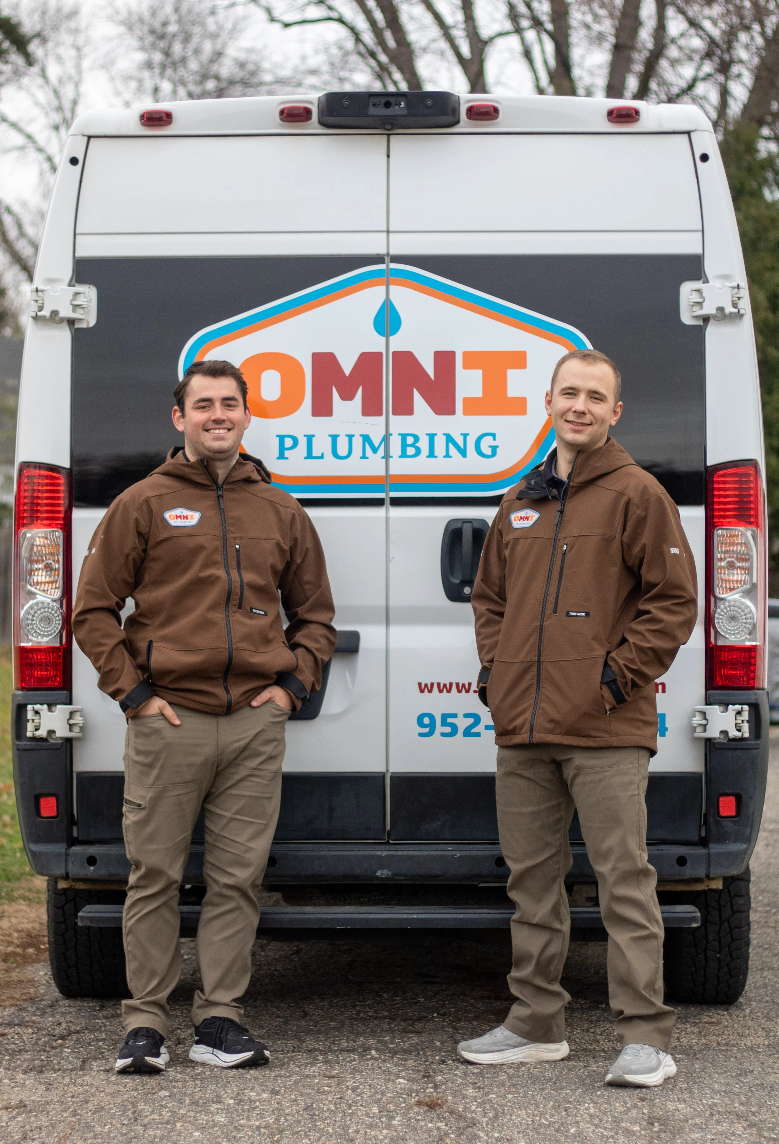 Two male service technicians standing in front of a white OMNI plumbing service van, both wearing brown jackets and khaki pants, smiling for the camera.