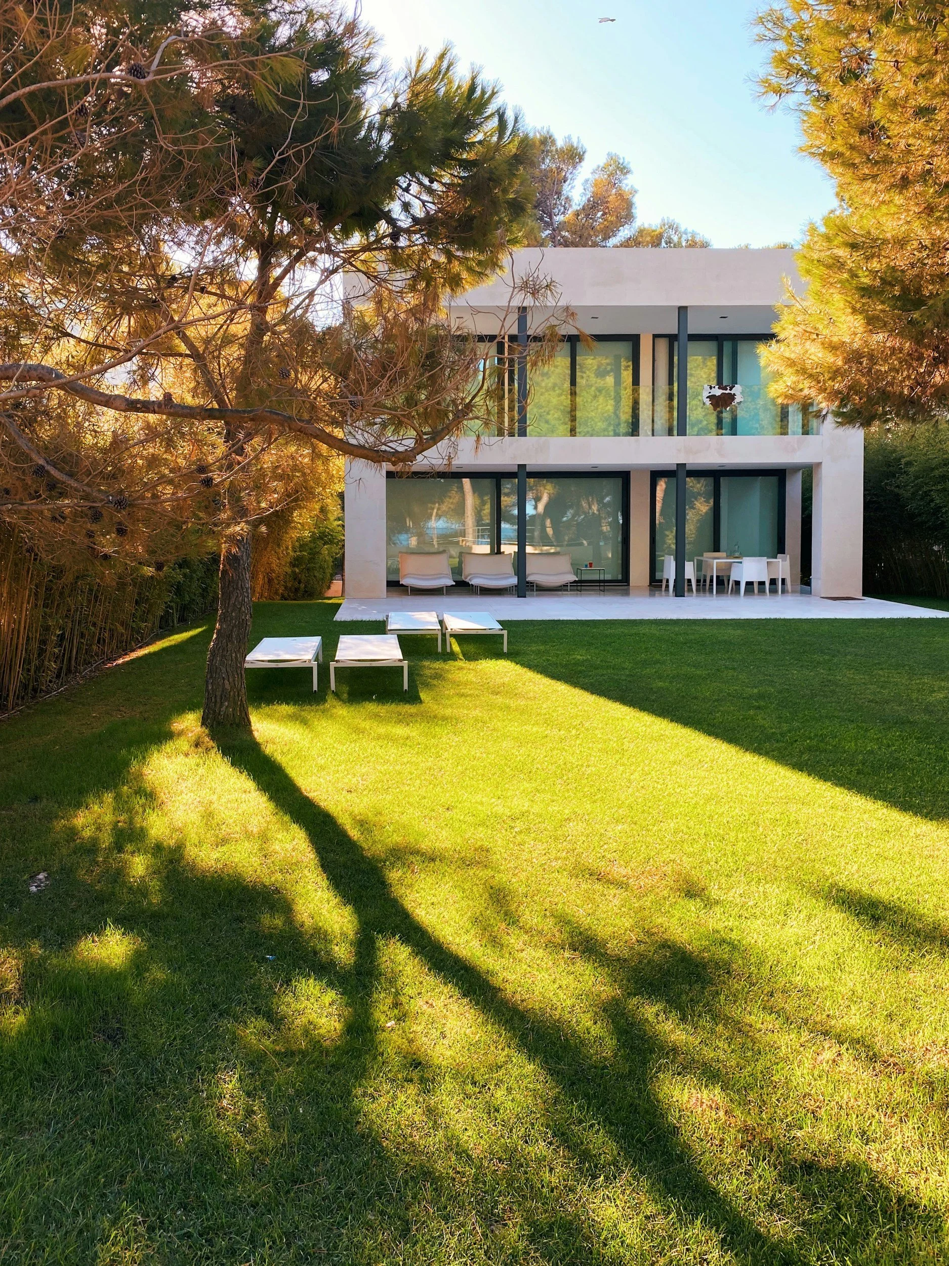 Modern two-story house with large glass windows and balconies, surrounded by trees and a grassy lawn, featuring outdoor furniture.
