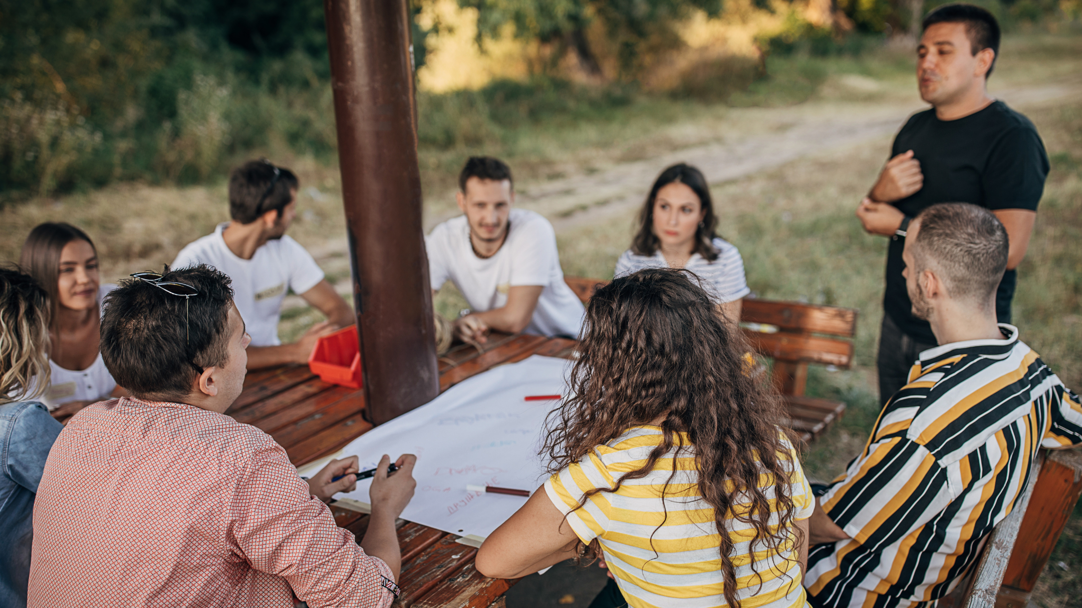 A group of people gathered around a wooden table outdoors, participating in a discussion or meeting, with a facilitator standing nearby.