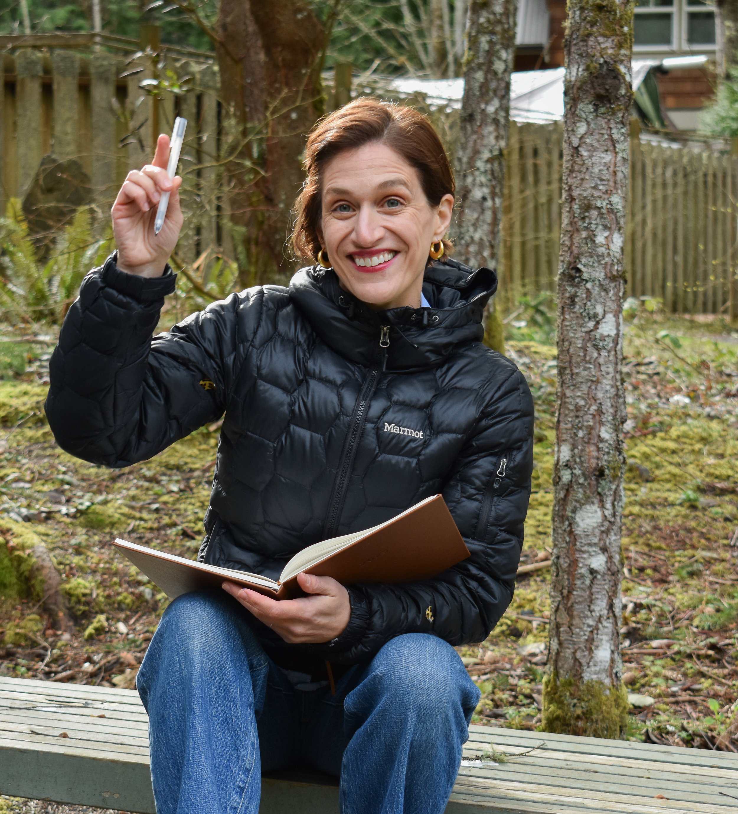 A woman sitting on a wooden bench outdoors with trees in the background, smiling, holding a pen in one hand and a notebook in her lap.