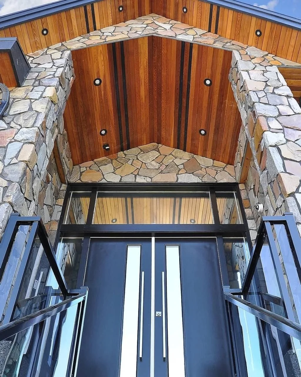 The image shows an upward view of a modern building entrance with glass doors, a stone archway and stone walls, and a wooden ceiling with recessed lighting.