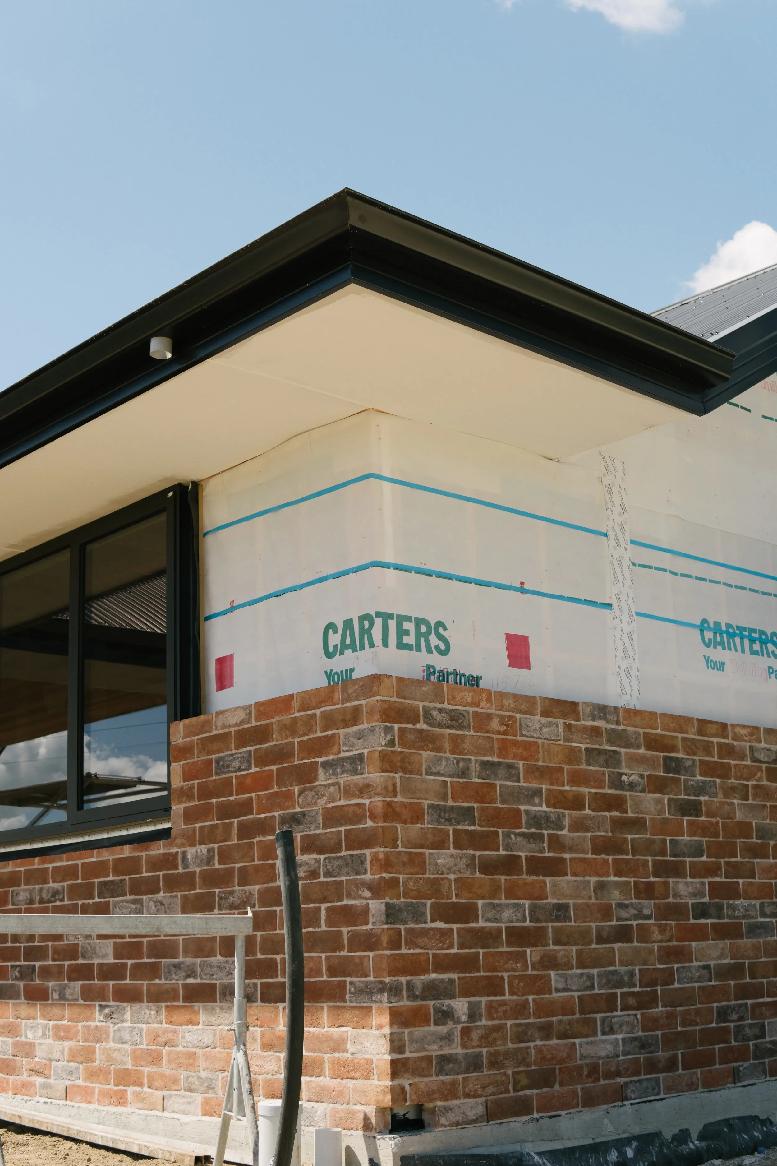 Part of a building under construction with brick walls, a window, and partially covered exterior wall with construction wrap showing the brand 'Carter's.' The roof has dark trim, and there are construction poles in front.