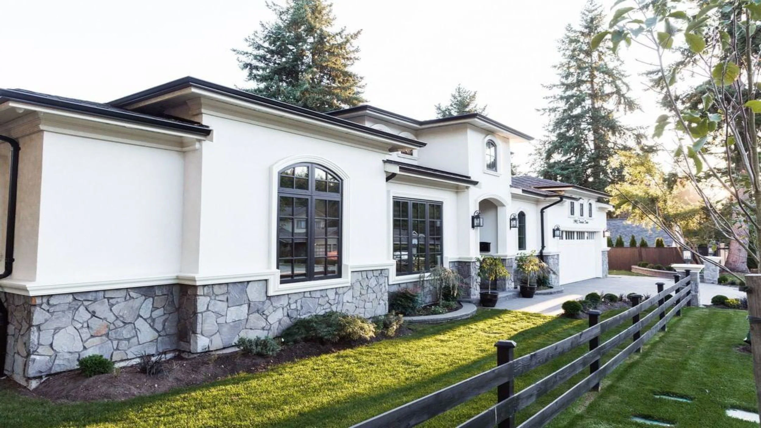 A modern white house with stone foundation, large windows, and a front yard with green grass, small shrubs, and a wooden fence.