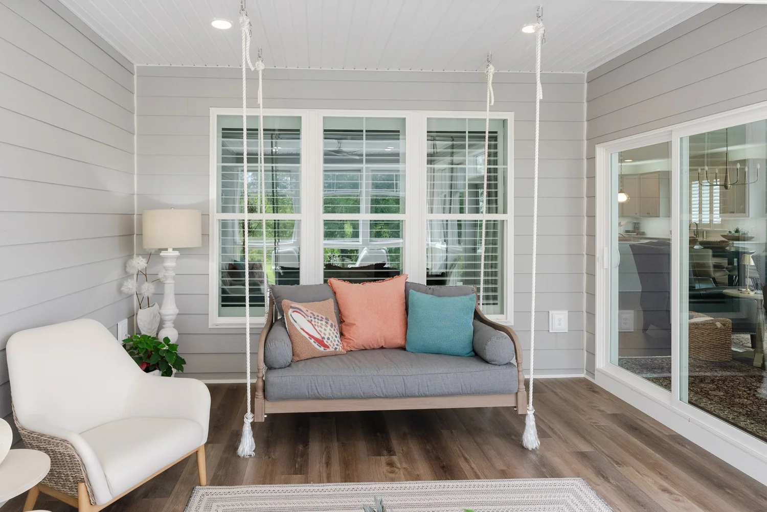 Rope-hung porch swing with gray cushions and coral pillows beneath the windows