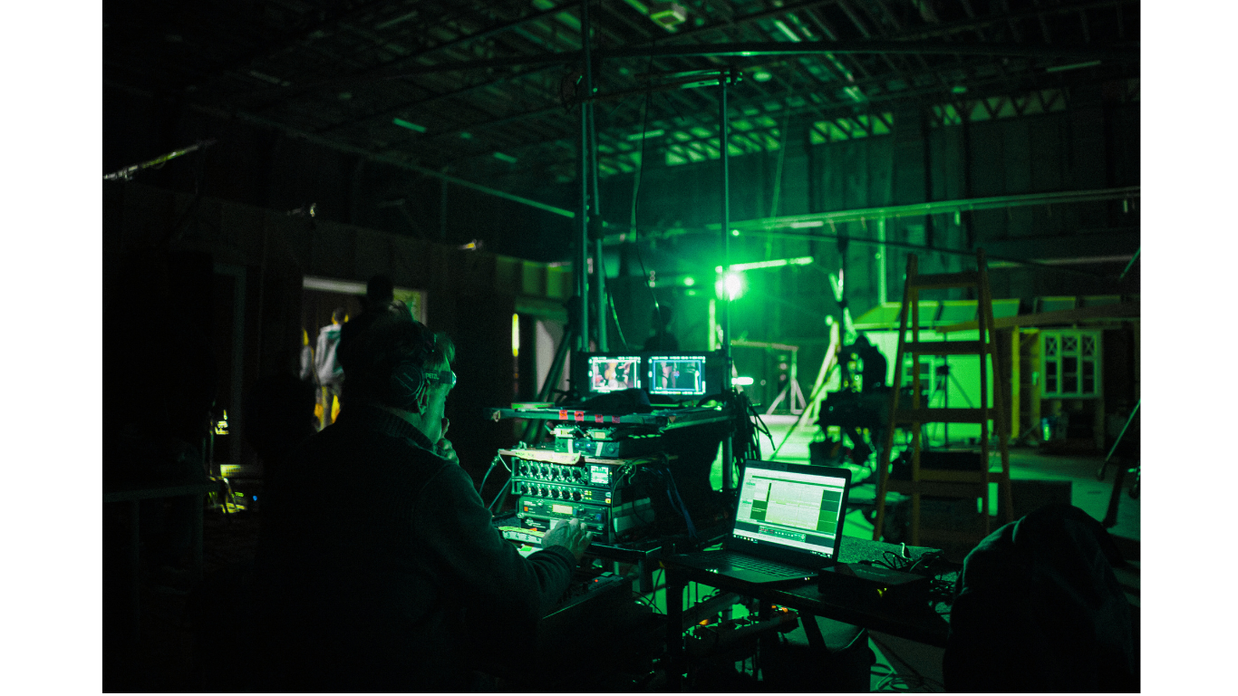 Behind the scenes at a photography or filming shoot with a green light in a warehouse or studio, featuring a technician working on sound or lighting equipment, and monitors showing footage.