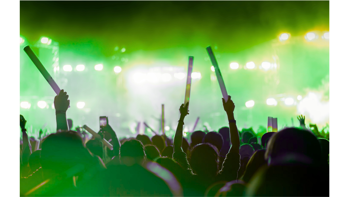 Crowd at concert waving glow sticks in front of a brightly lit stage with green and white lights.