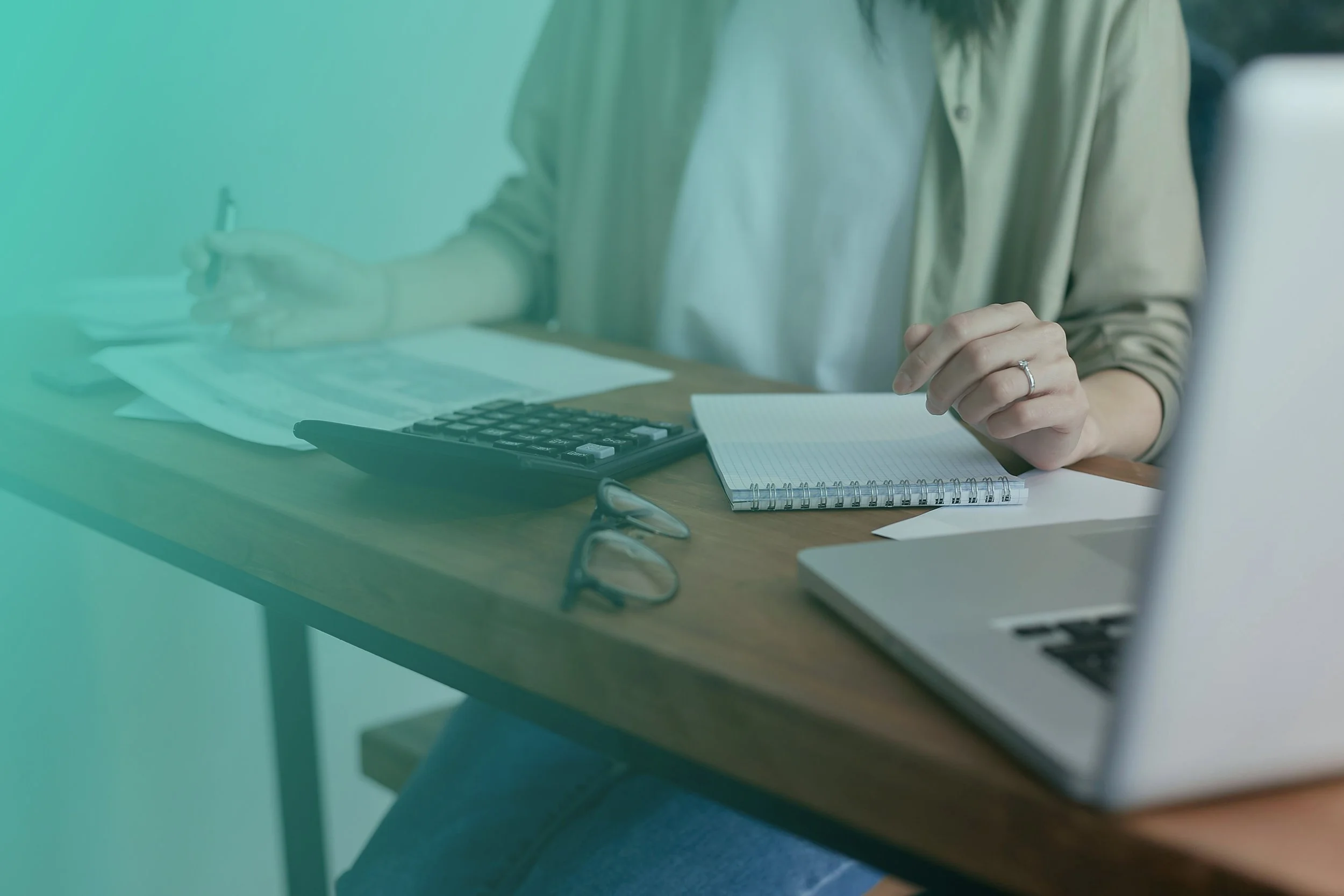 A woman working at a desk with a laptop, calculator, notebook, glasses, and papers