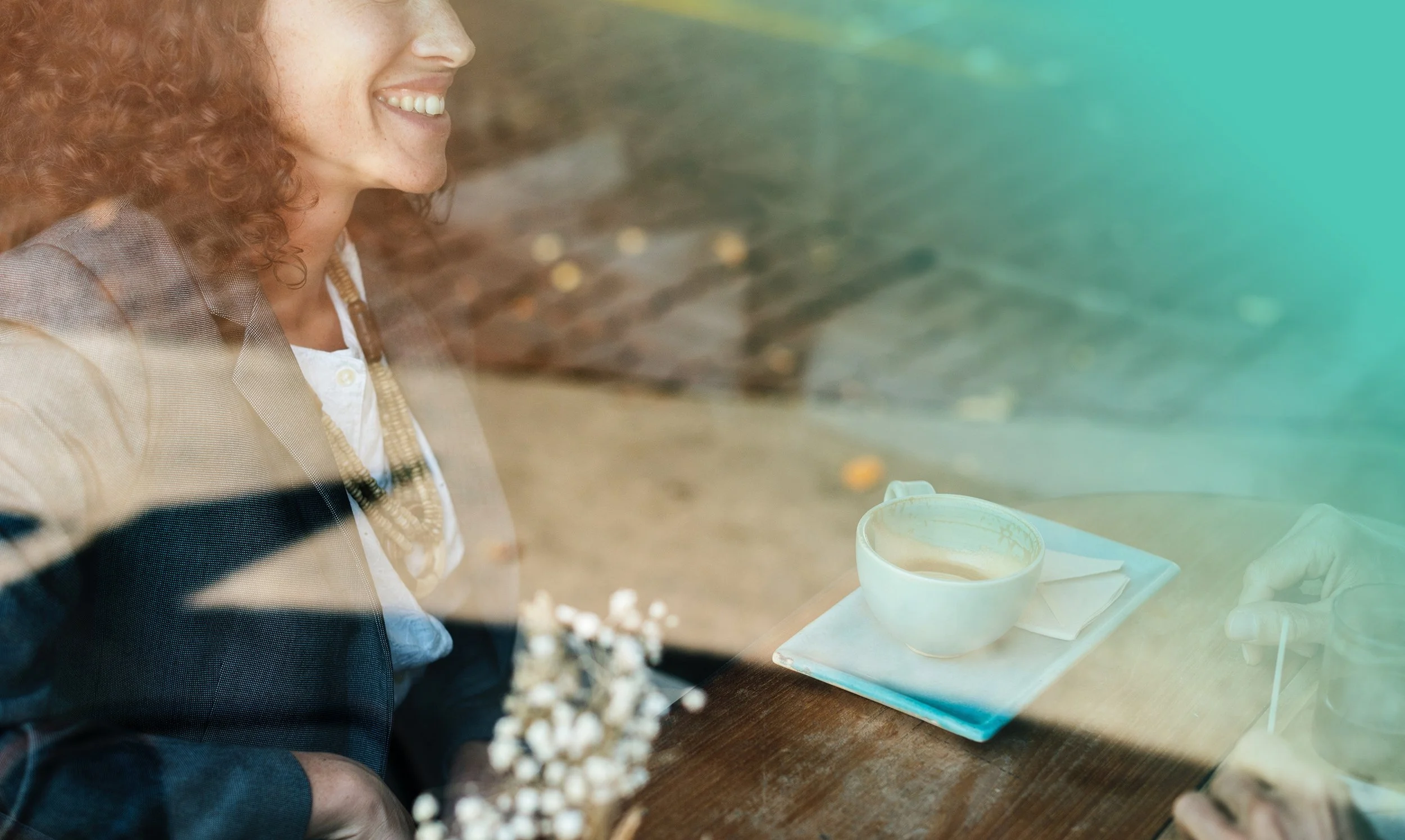 A woman smiling at a cafe table with a cup of coffee and a hand holding a cellphone through a window reflection.