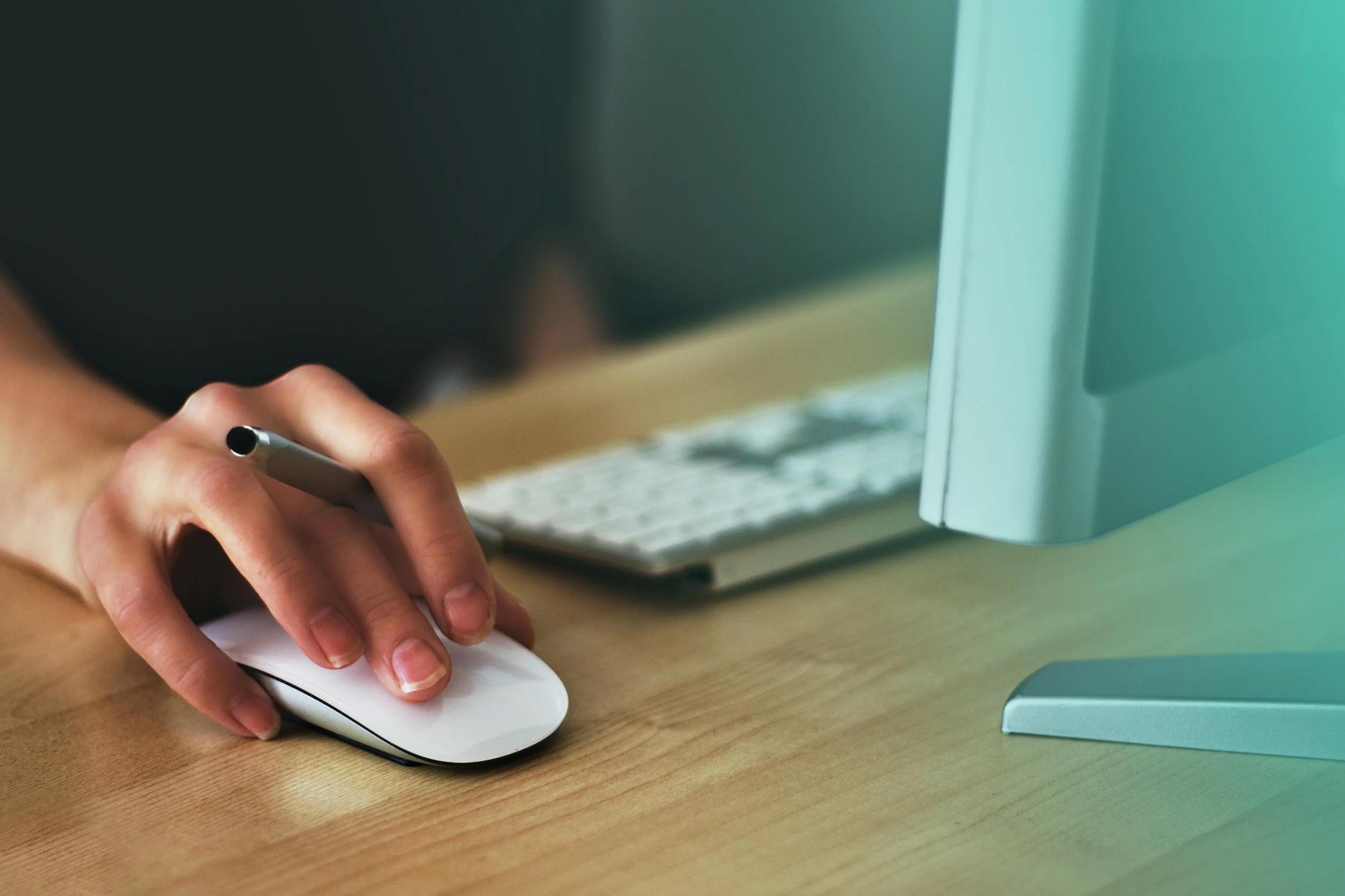 Close-up of a person's hand with short nails operating a white computer mouse on a wooden desk, with a keyboard and a large monitor in the background.