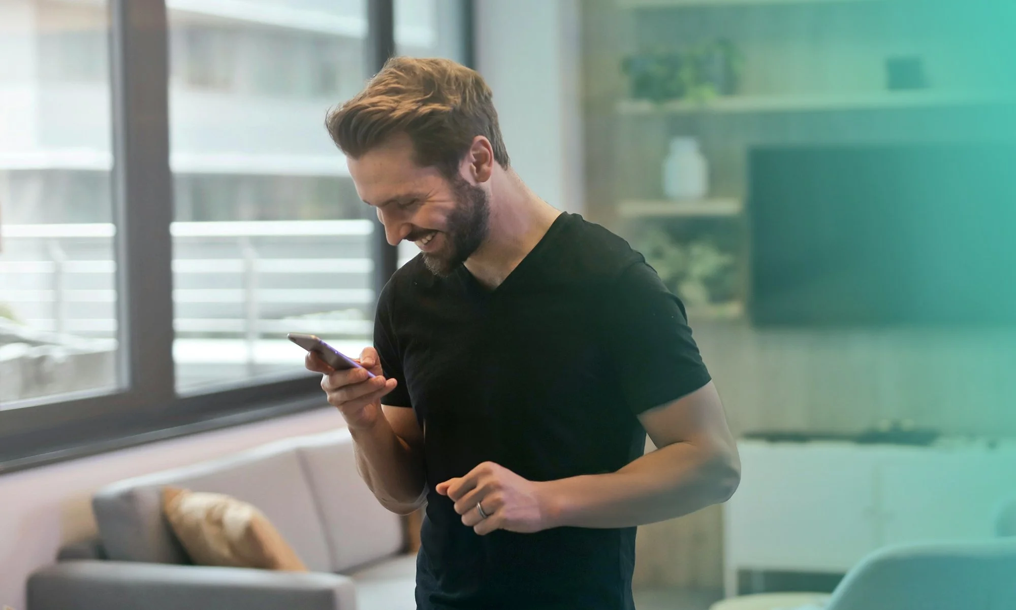 A young man with a beard and short hair, wearing a black t-shirt, happily looking at his smartphone while standing in a bright room with large windows and a sofa with cushions and television in the background.