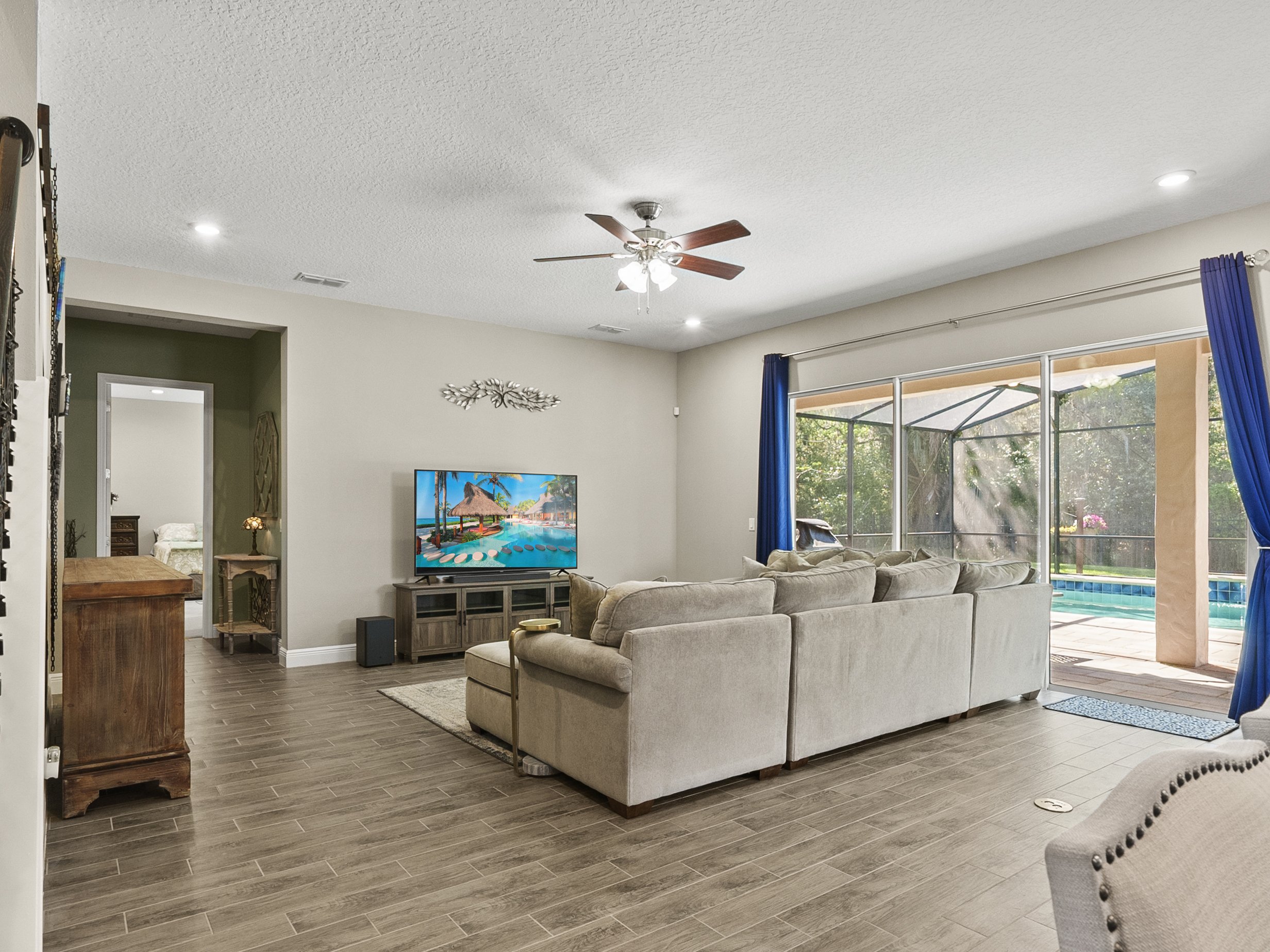 Modern living room with beige sectional sofa, hardwood floors, ceiling fan, large TV, and glass doors leading to a pool area with blue curtains.
