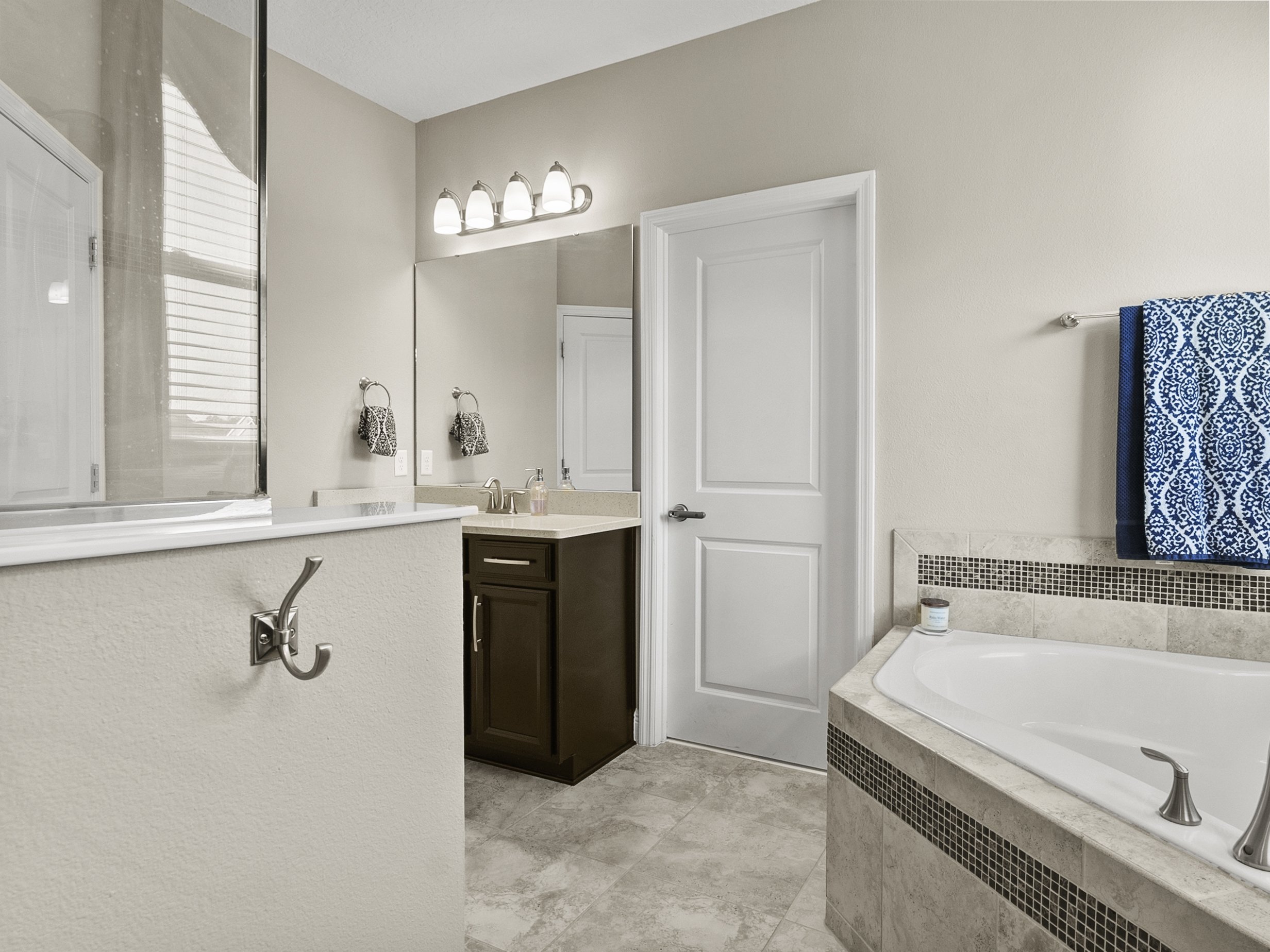 Modern bathroom with bathtub, vanity, and towel rack. Features a large mirror, overhead lights, and a blue patterned towel. Neutral-toned tiles and wall paint.