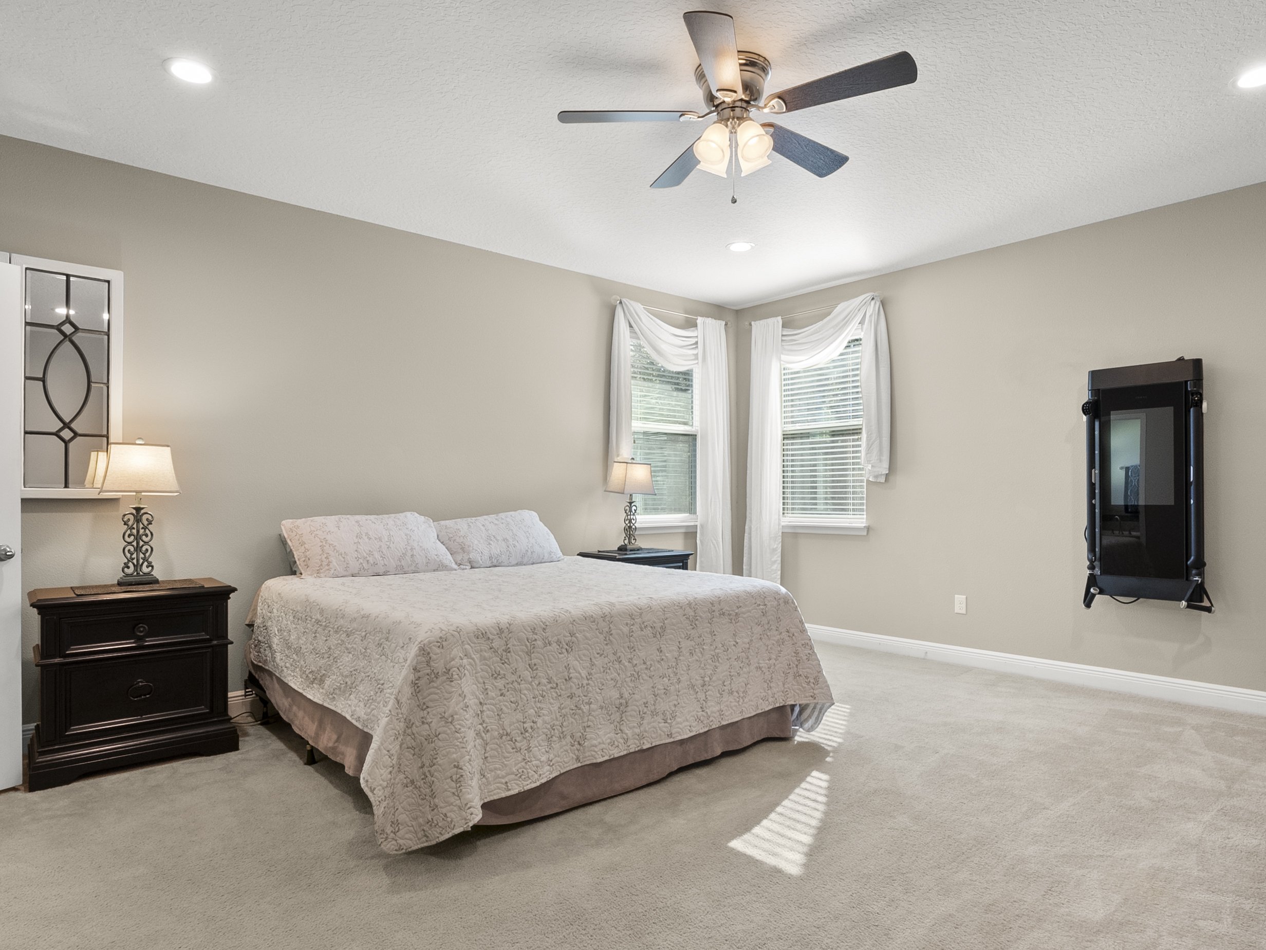 Spacious bedroom interior with a king-size bed, beige walls, and carpet. The room features a ceiling fan with lights, wall-mounted workout equipment, and two nightstands each with a lamp. Natural light streams in through two windows with white drapes