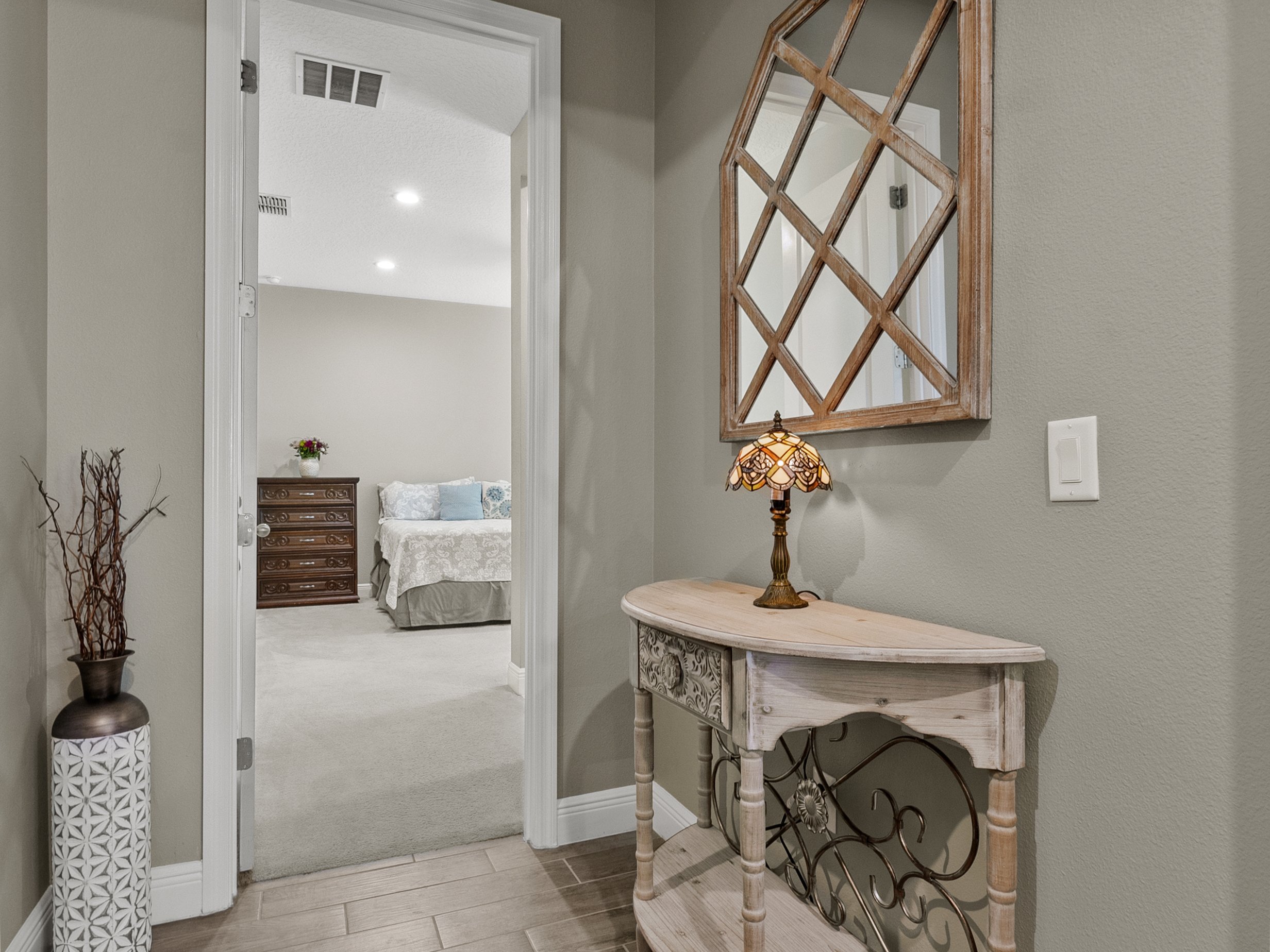Hallway with small table and lamp, decorative mirror, and vase with sticks, leading to bedroom with bed and dresser.