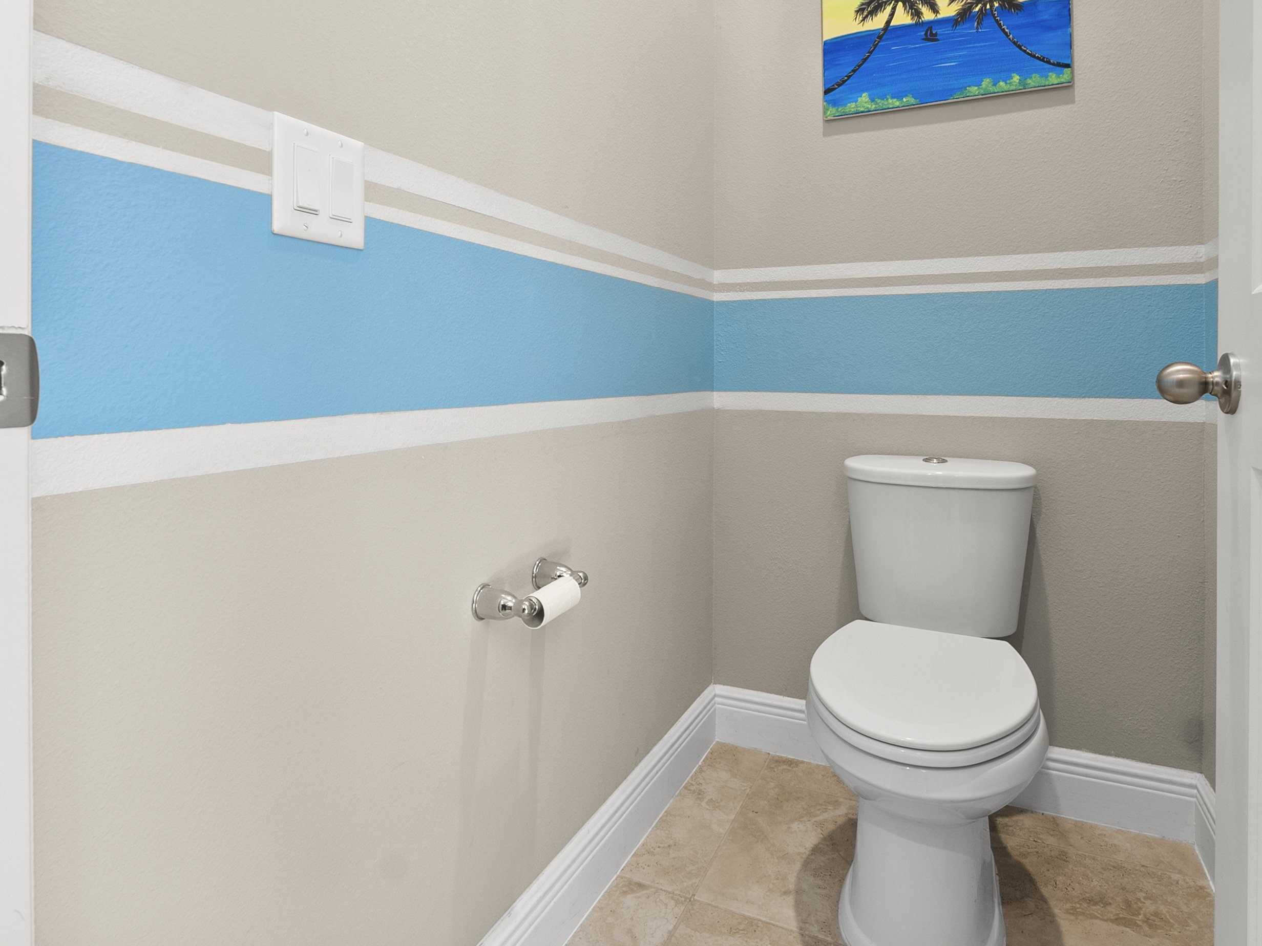Bathroom with white toilet, beige walls with a blue stripe, white light switch, and a roll of toilet paper on a holder; a painting of a beach scene on the wall.