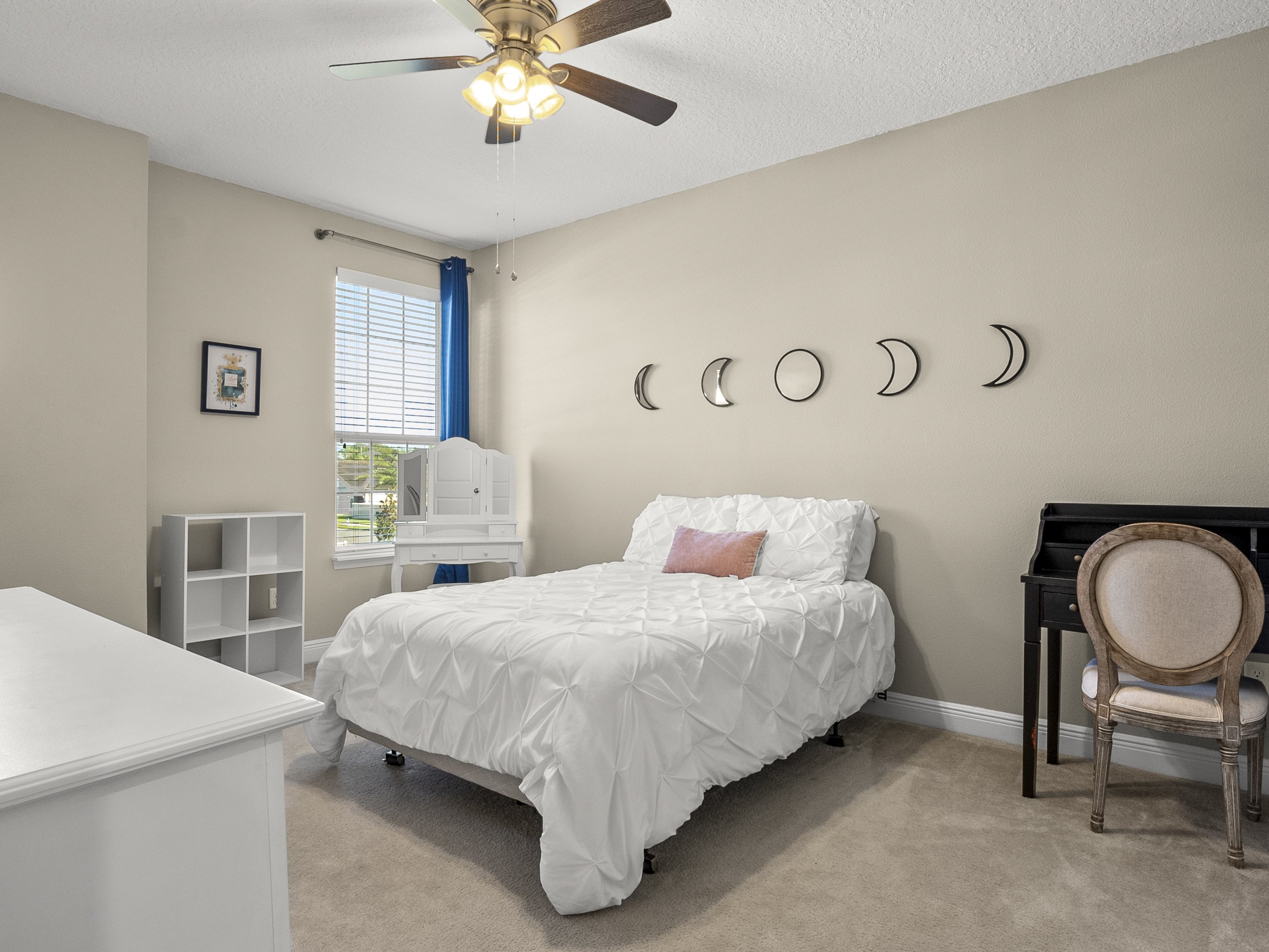 Minimalist bedroom with a white bed and moon phase wall decor, featuring a ceiling fan, a window with blue curtains, a white vanity desk, and a black writing desk with a chair.