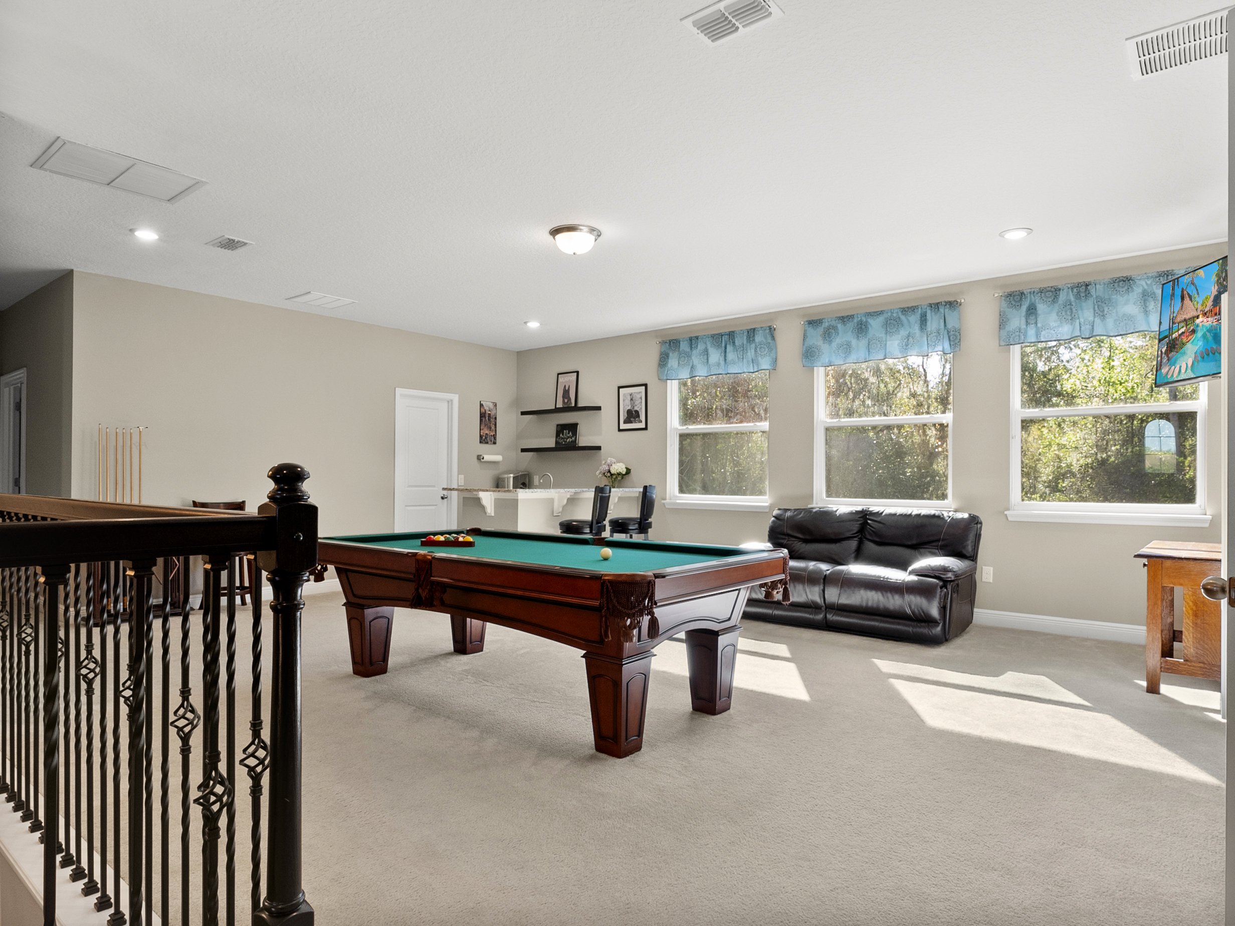 Spacious game room featuring a pool table, black leather sofa, large windows with blue curtains, and an entertainment area with cues on a stand, a desk, and a TV mounted on the wall.