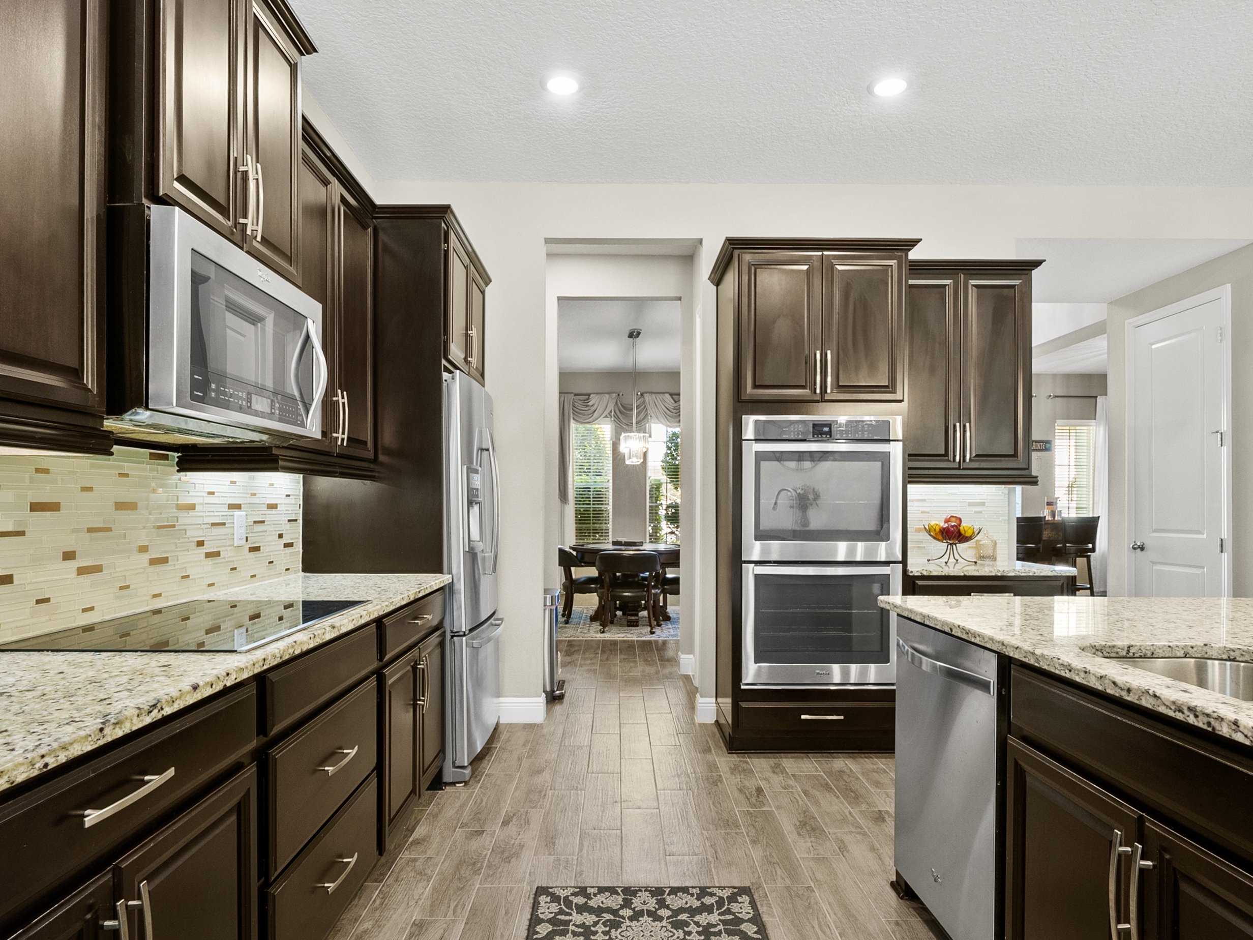 Modern kitchen with dark wood cabinets, stainless steel appliances, granite countertops, and a tile backsplash. An open doorway leads to a dining area in the background.
