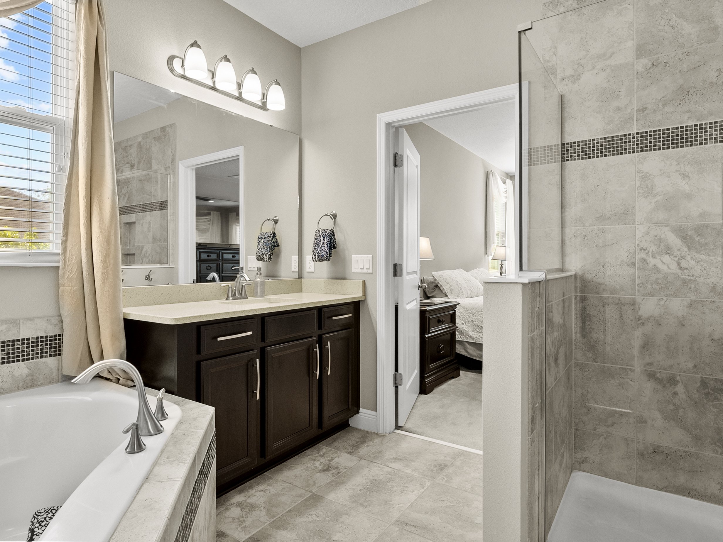 Modern bathroom with a soaking tub, double sink vanity with dark wood cabinets, and beige tile shower. The room is well-lit with natural light from a window and has a doorway leading to a bedroom.