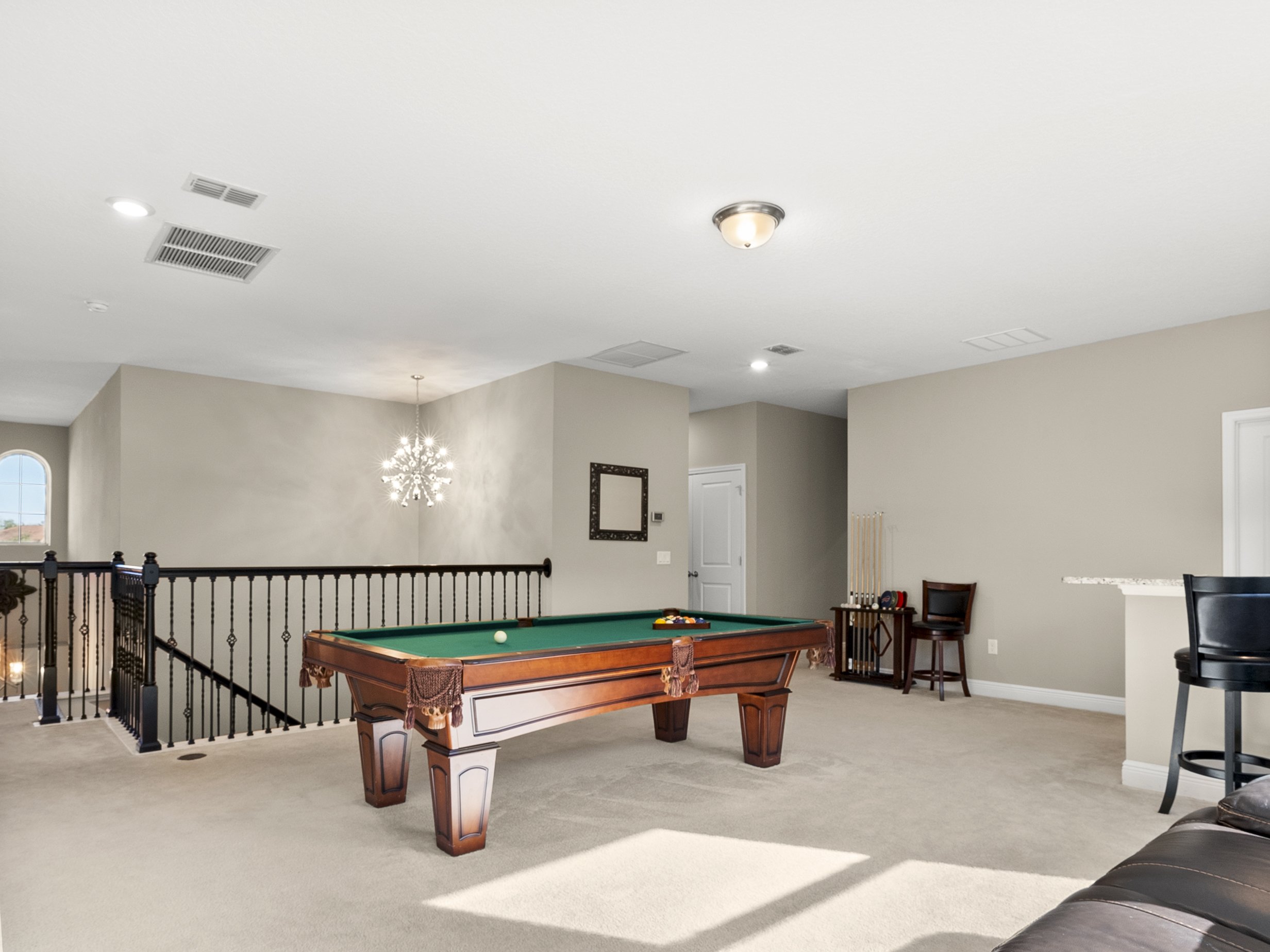 Room with a pool table, carpeted floor, elegant lighting fixture, and bar stools near a railing.
