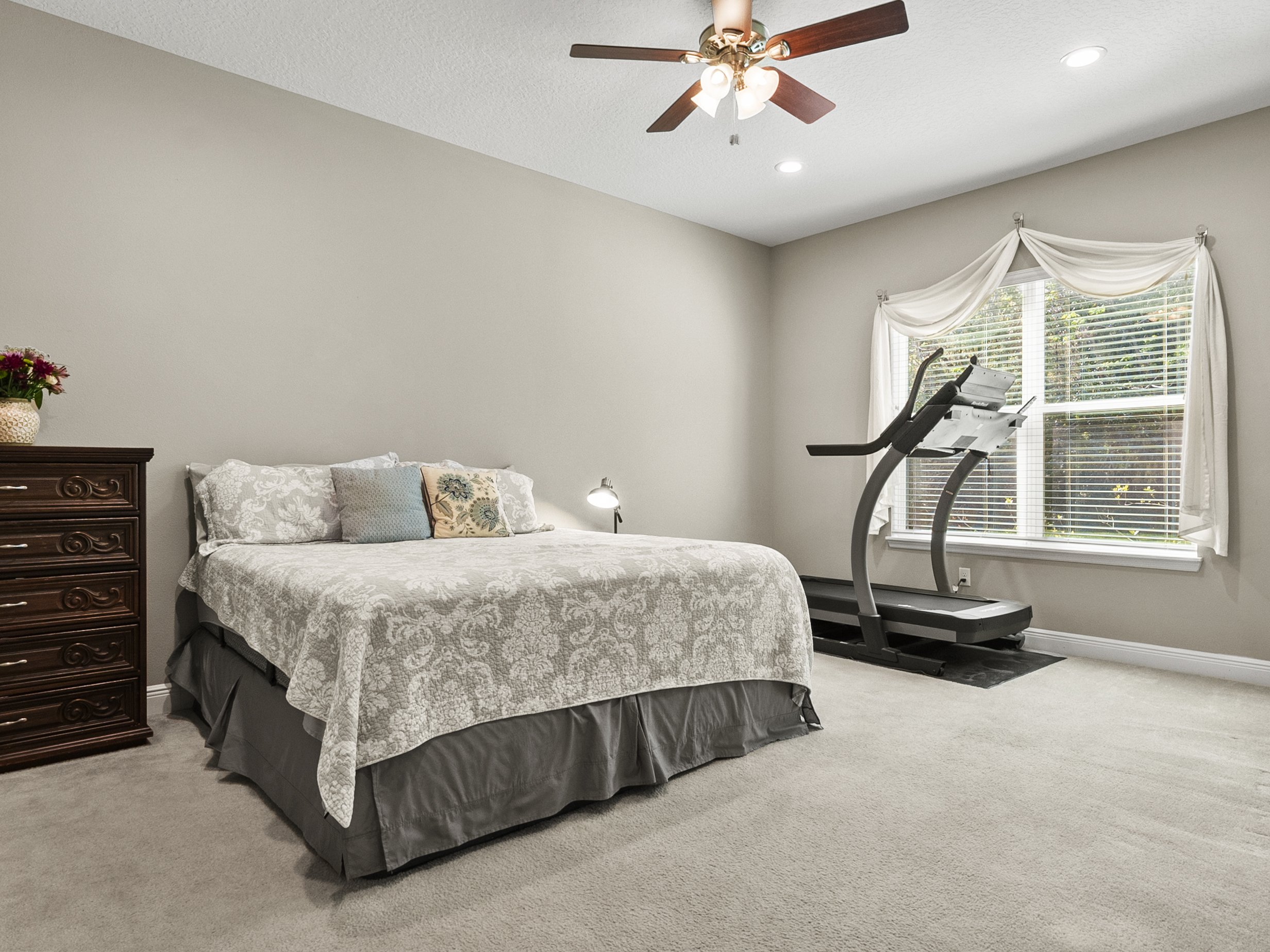 Bedroom with bed, treadmill, dresser, ceiling fan, and window with drapes.