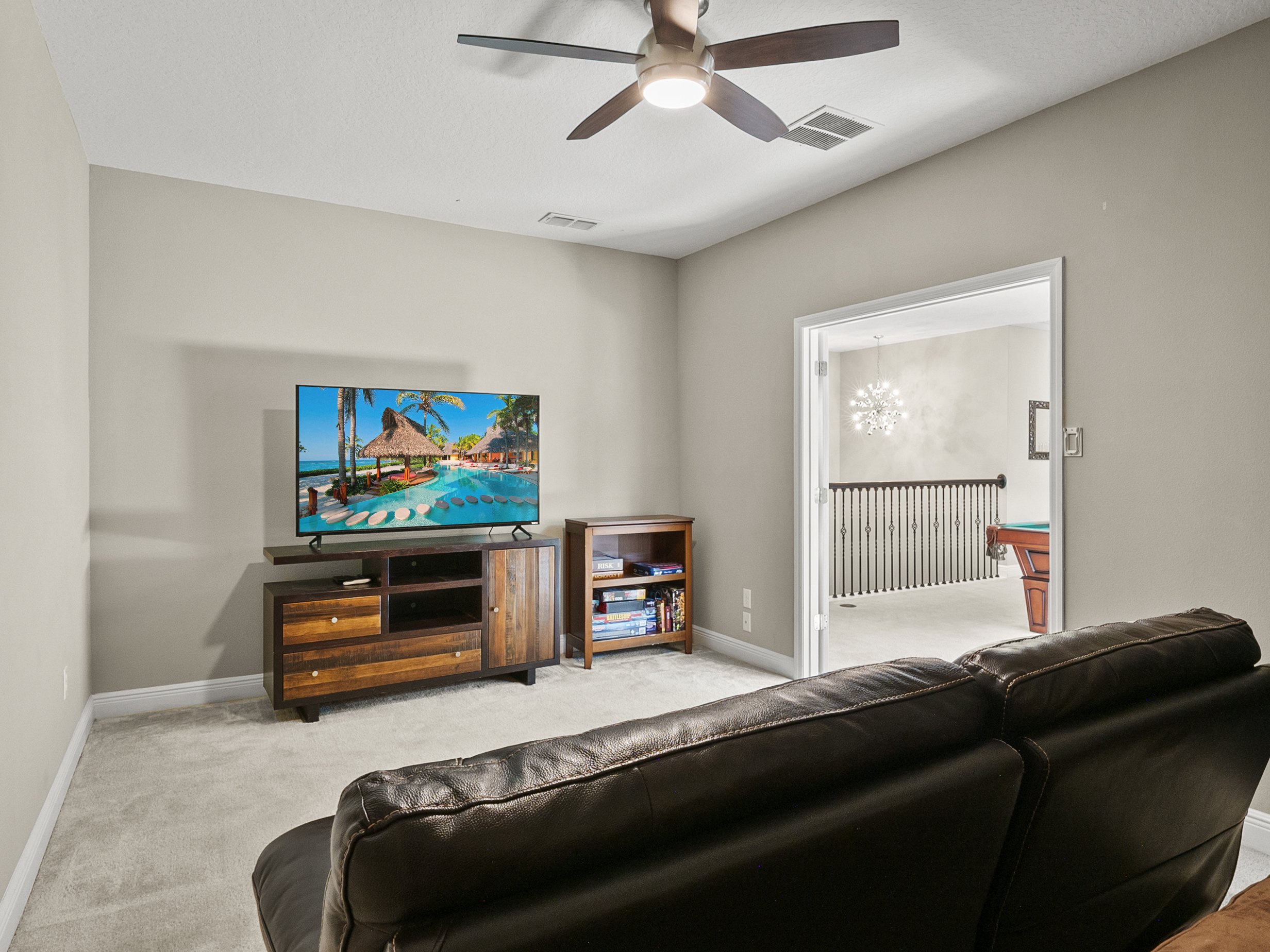 Living room with a brown leather couch facing a flat-screen TV on a wooden stand, displaying a tropical beach scene, with an open doorway leading to a room with a billiards table.