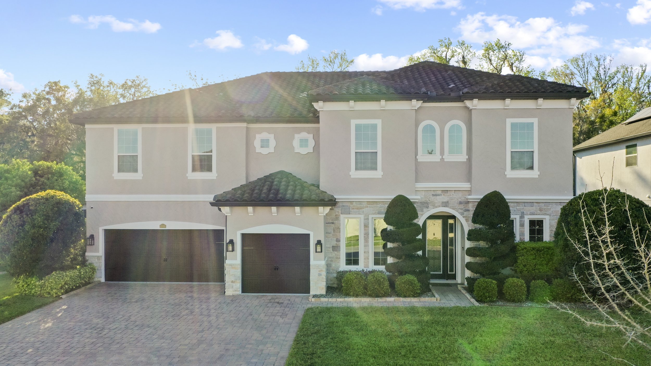 Two-story modern house with stucco exterior, double garage doors, decorative plants, and paved driveway under a clear blue sky.