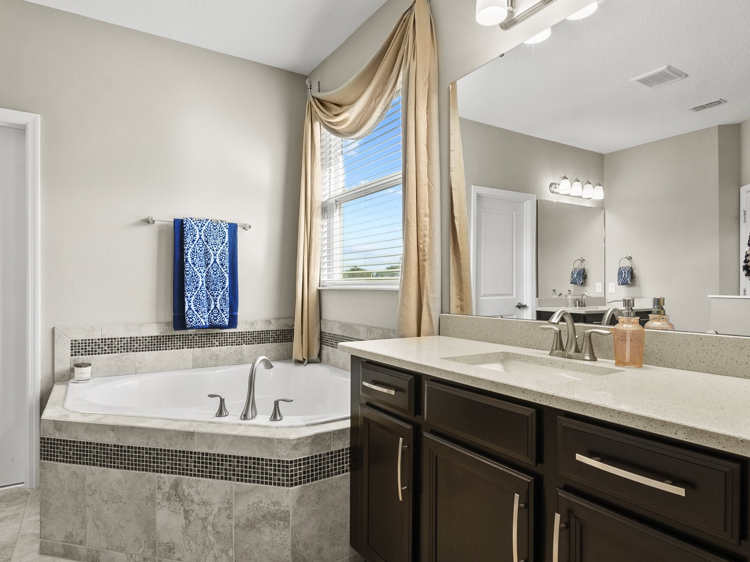 Modern bathroom with a large corner bathtub, dark wood vanity, a wide mirror, and a window with beige curtains. The bathtub is adorned with a blue patterned towel. The countertop has dual faucets and is decorated with soap dispensers.