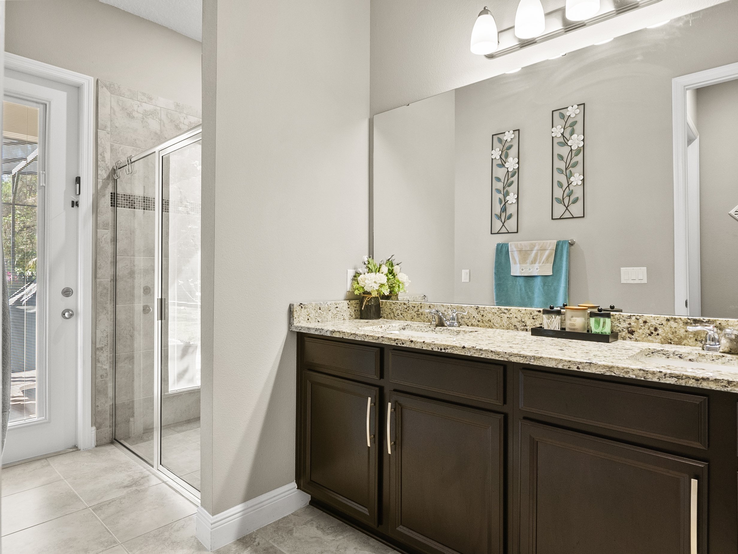 Modern bathroom with dual sink vanity, granite countertop, glass shower, and decorative wall art. Tiled floor and bright lighting.
