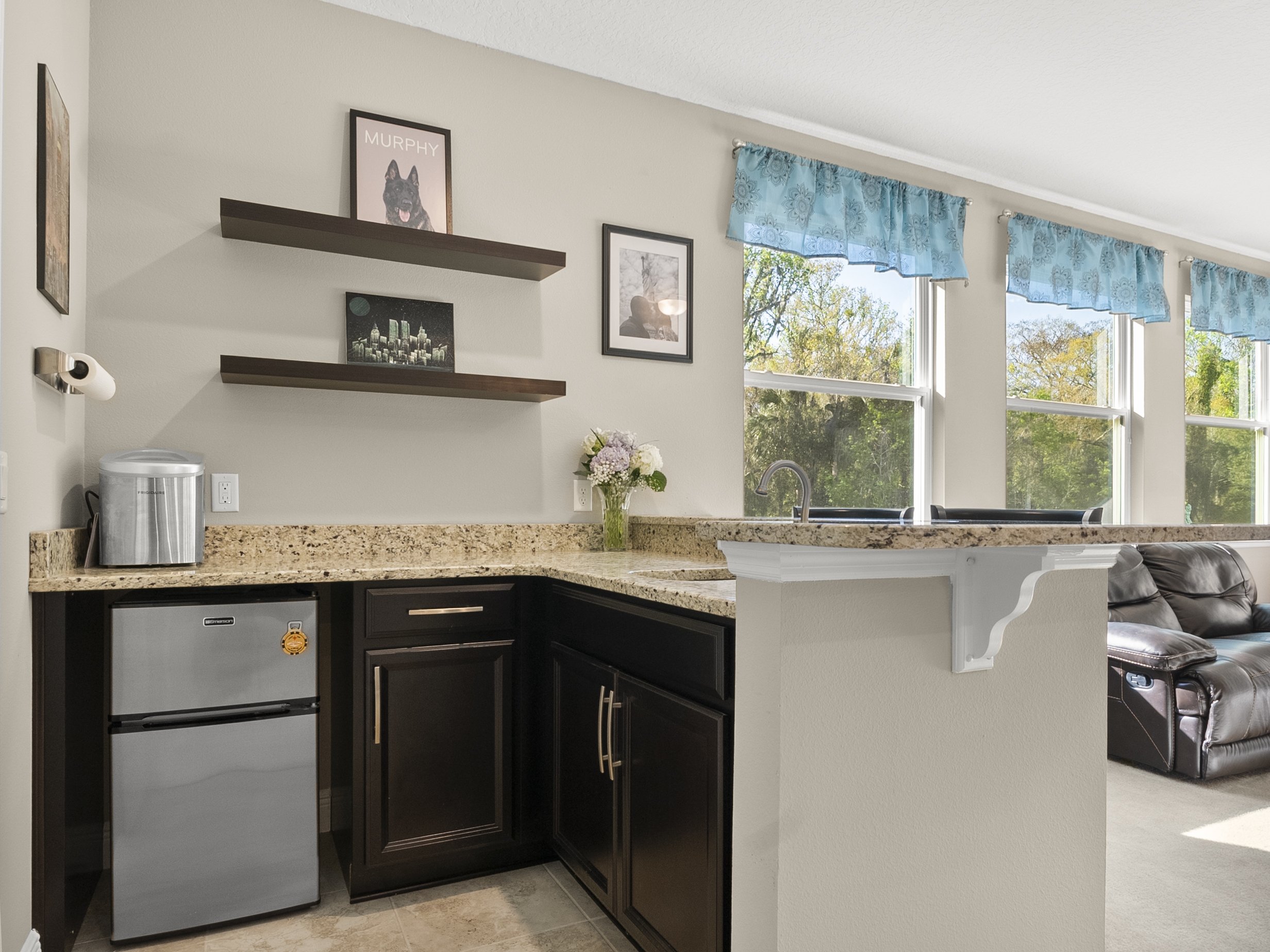 Interior of a modern kitchen with a small refrigerator, granite countertops, dark wood cabinets, and shelving. There is a decorative picture on the wall, a vase with flowers on the counter, and multiple windows with blue curtains in the background.