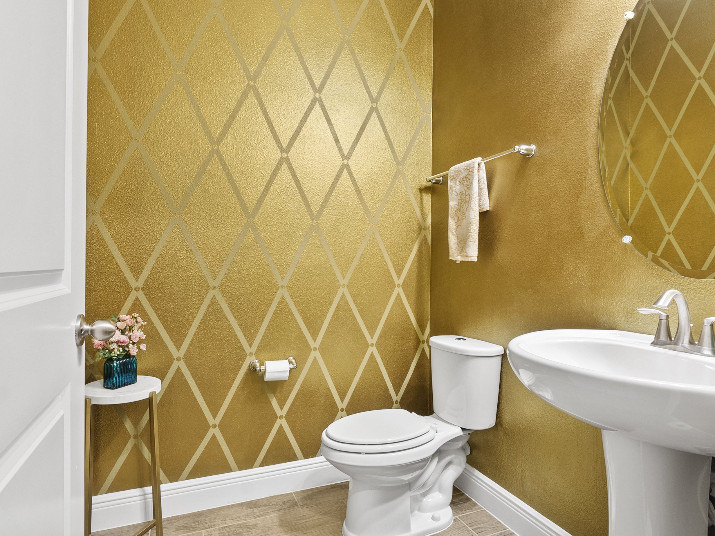 Luxury bathroom with gold diamond-patterned walls, white toilet, pedestal sink, oval mirror, towel on a rack, small table with flowers, and wood-textured floor tiles.