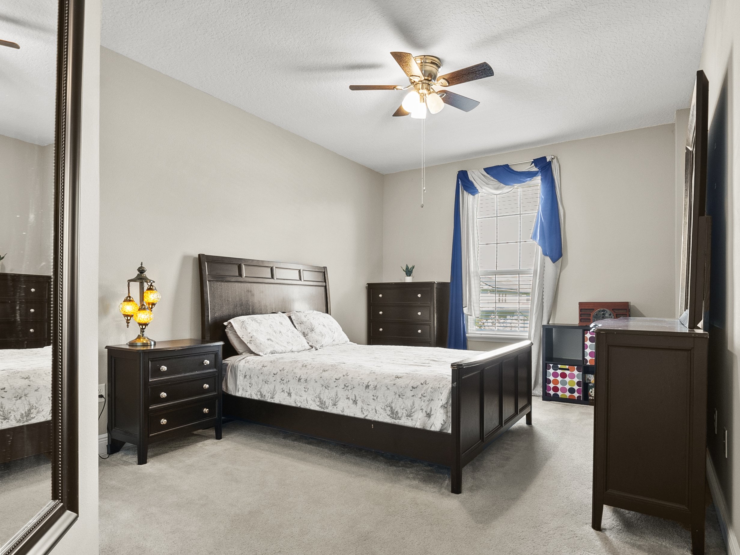 A bedroom with beige walls featuring a dark wood bed with floral bedding, a matching nightstand with a vintage lamp, a dresser, and a window with blue and white curtains.