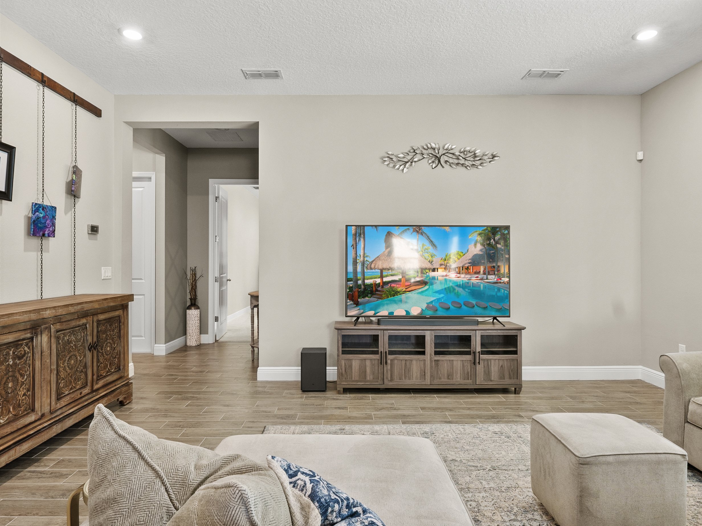 Living room with decorative wooden cabinet, wall art, and a flat-screen TV displaying a tropical resort scene. Modern furnishings and neutral decor.